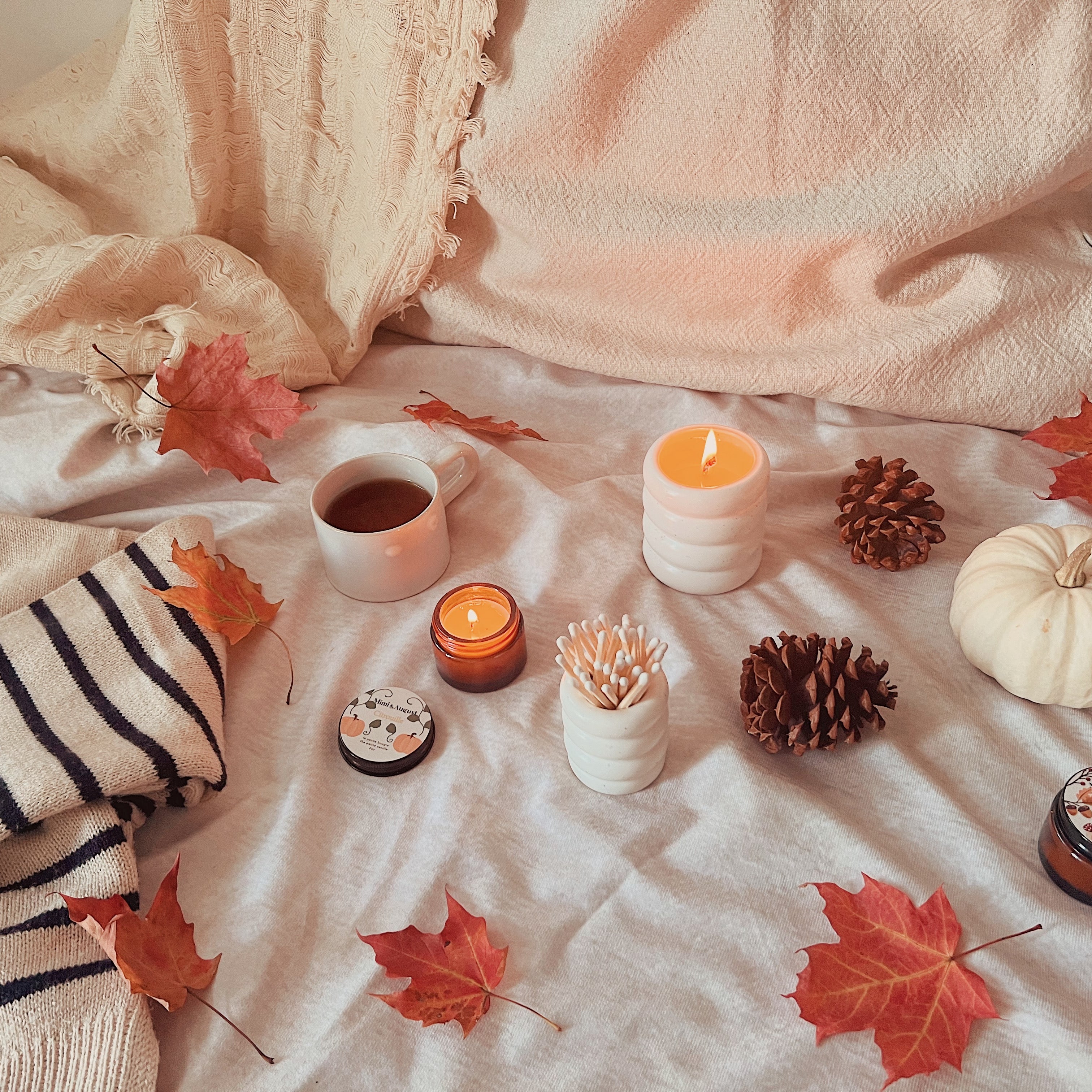 Mimi & August exlusive fall candles sitting on a warm cozy fall bed beside a sweater, a blanket, a pillow, a warm cup of tea, fall leaves and a white pumkin