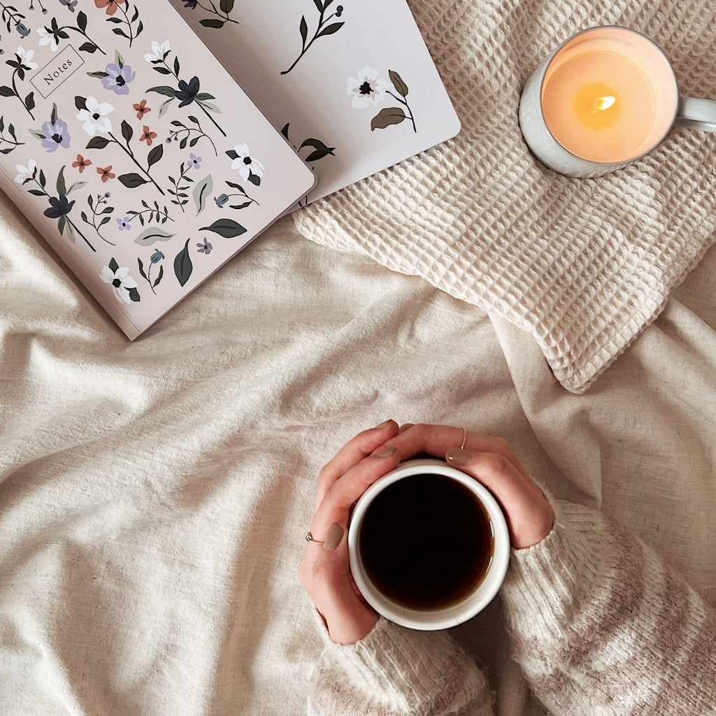 Hands holding a cup of tea on a bed beside notebooks and Botanica candle from Mimi & August