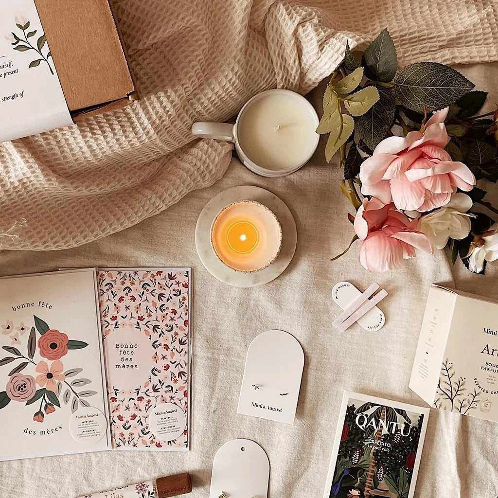 Mother's Day gift ideas featuring floral-themed greeting cards, a lit soy candle in a ceramic cup, artificial pink and white flowers, and a beige gift box
