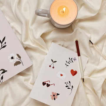 Floral notebook, Valentine's Day card and mug scented candle from Mimi & August laid on a cozy blanket