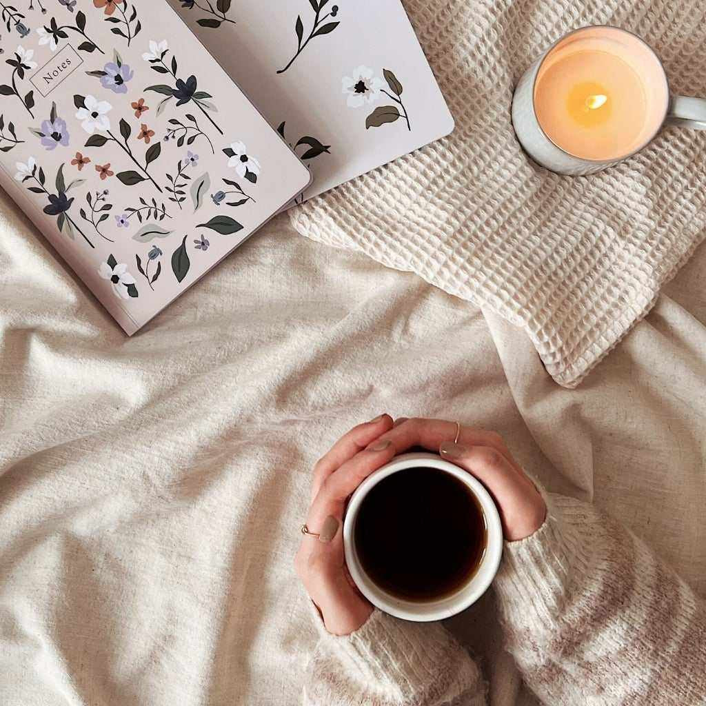 Hands holding a white ceramic mug filled with black coffee on a beige fabric surface with a lit candle and floral notebook for winter self-care tips