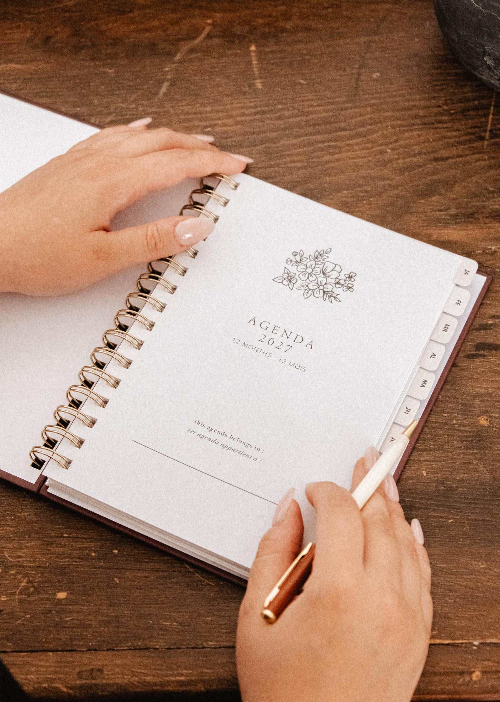 A person opens a spiral-bound Wildflowers – 2027 agenda by Mimi & August on a wooden table, holding a pen and turning the page with manicured hands—perfect for monthly and weekly planning.