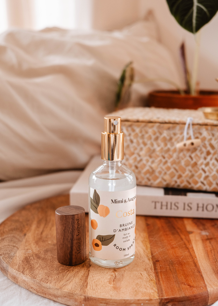 A bottle of Mimi & August Costa Room Spray with a floral design on a wooden tray next to a plant and books with the title "This is Home.