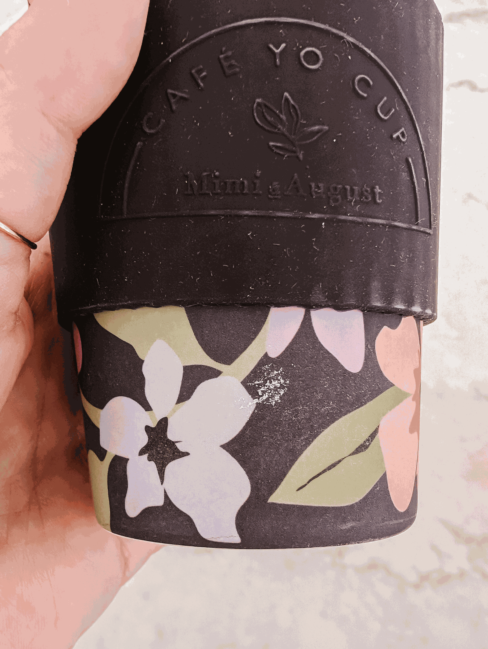 A person holding up a Defect Café Yo Cup by Mimi & August with flowers and a silicone lid on it.