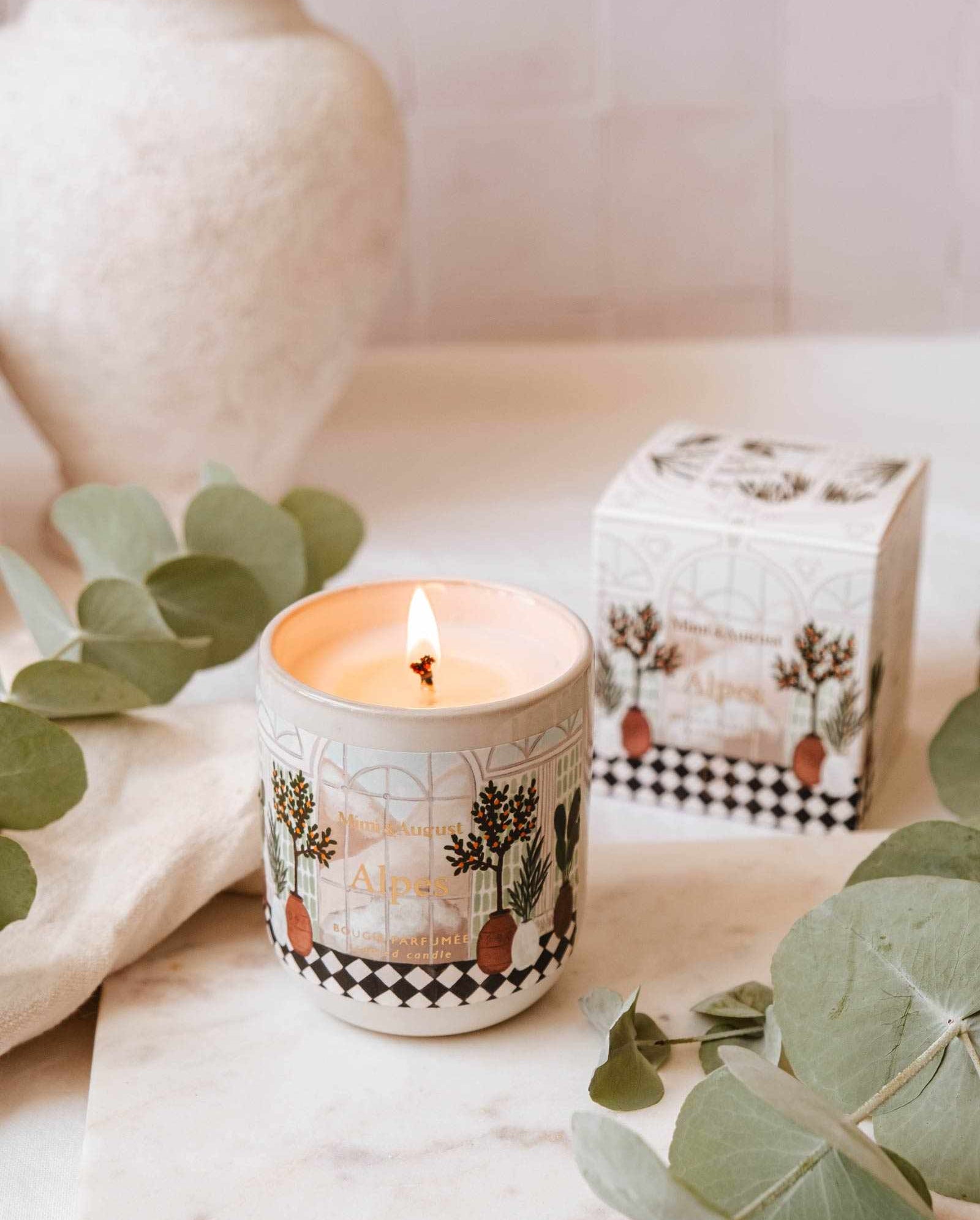 An Alpes - Reusable Candle by Mimi & August in a decorative ceramic container casts a warm glow on a marble surface near eucalyptus leaves, with its elegant reusable box positioned in the background.