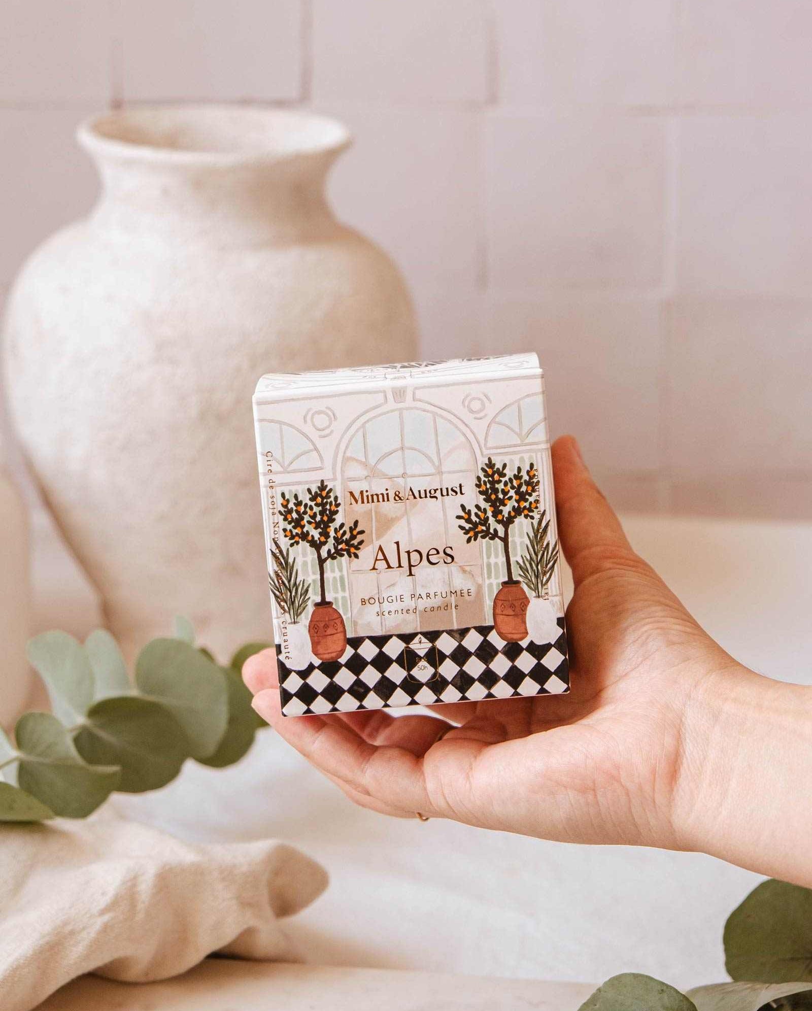 A hand holds a box of "Mimi & August Alpes - Reusable Candle" with an illustrated design. A white ceramic vase and green eucalyptus leaves are in the blurred background, hinting at the fresh scent and elegance of these reusable candles.