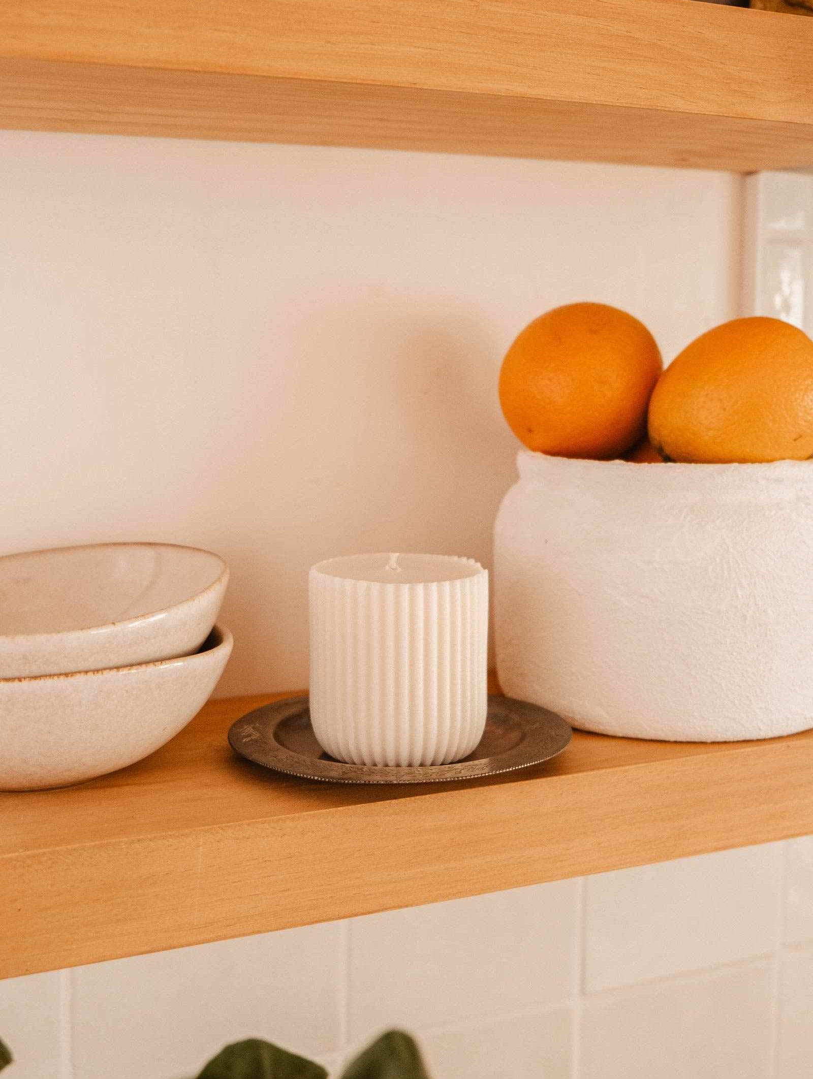 Three stacked ceramic bowls, a Mimi & August Candle Refill Cielo (soy wax, fig cassis amber scent) on a metal saucer, and a white container with oranges sit on a wooden shelf.