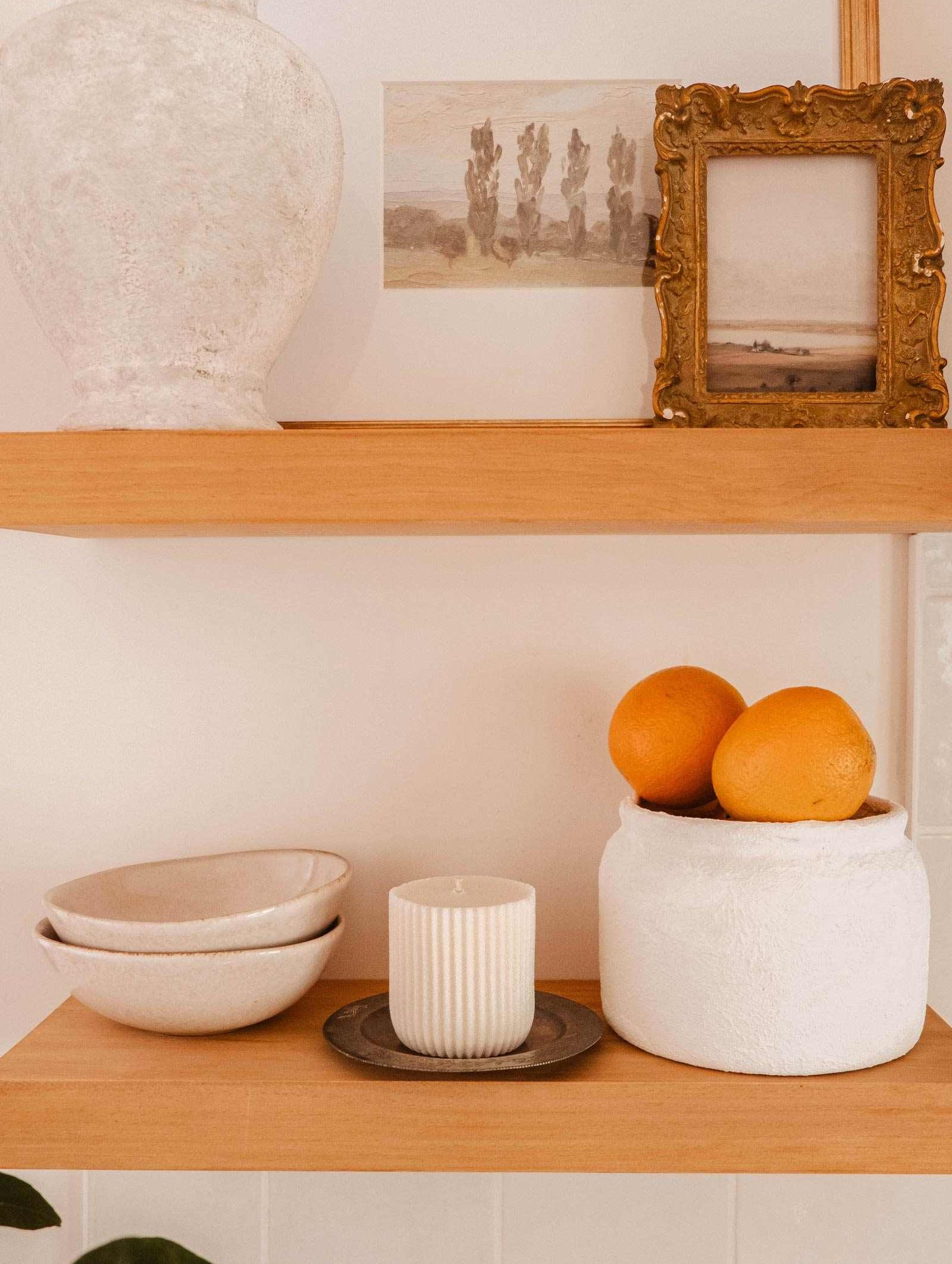 Wooden shelves display ceramic bowls, a Mimi & August Candle Refill Alpes, a white vase, framed landscape art, and a white container with oranges—each piece brings the cozy charm of an alpine fragrance.