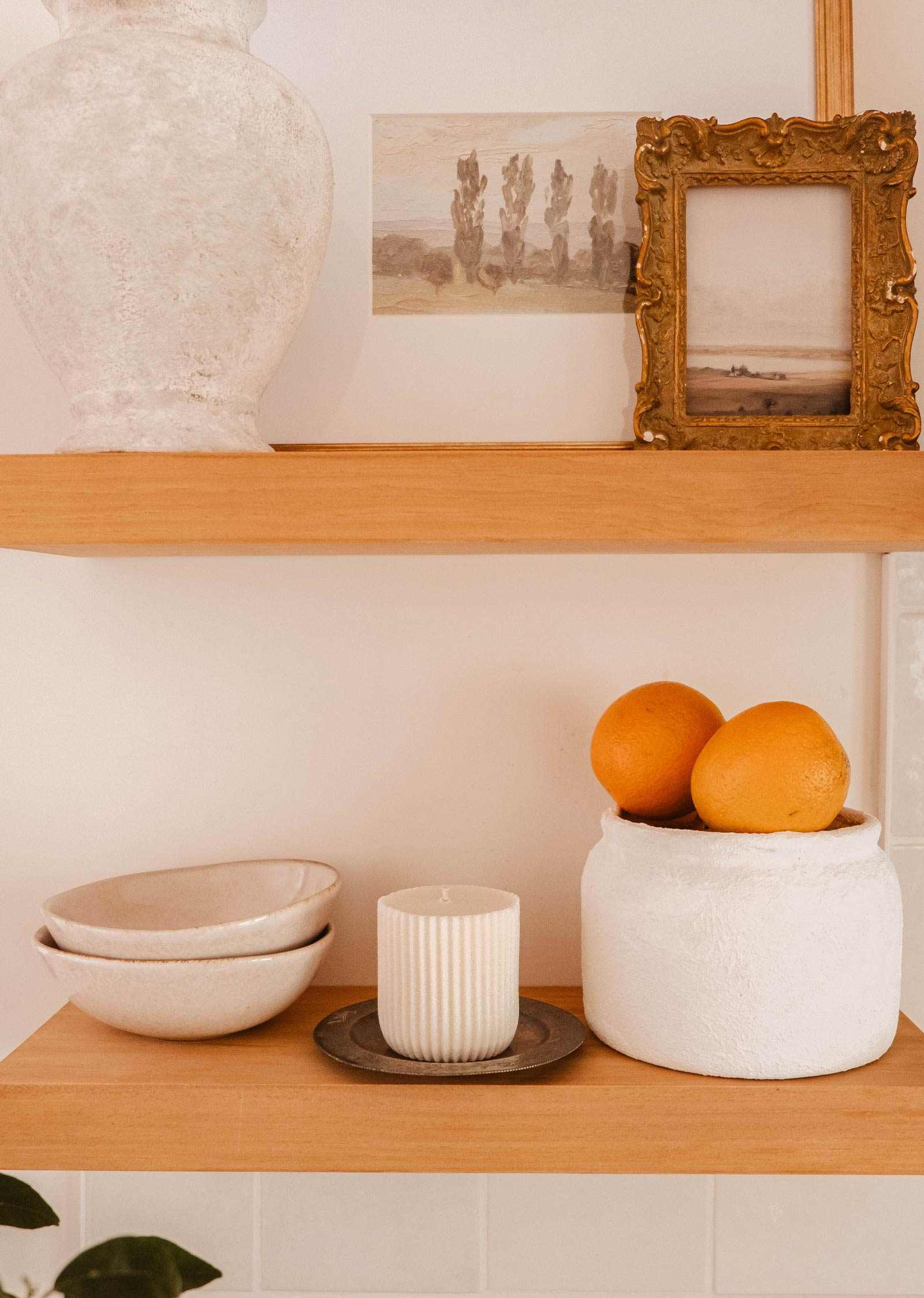 Wooden shelves display ceramic bowls, a Mimi & August Candle Refill Alpes, a white vase, framed landscape art, and a white container with oranges—each piece brings the cozy charm of an alpine fragrance.