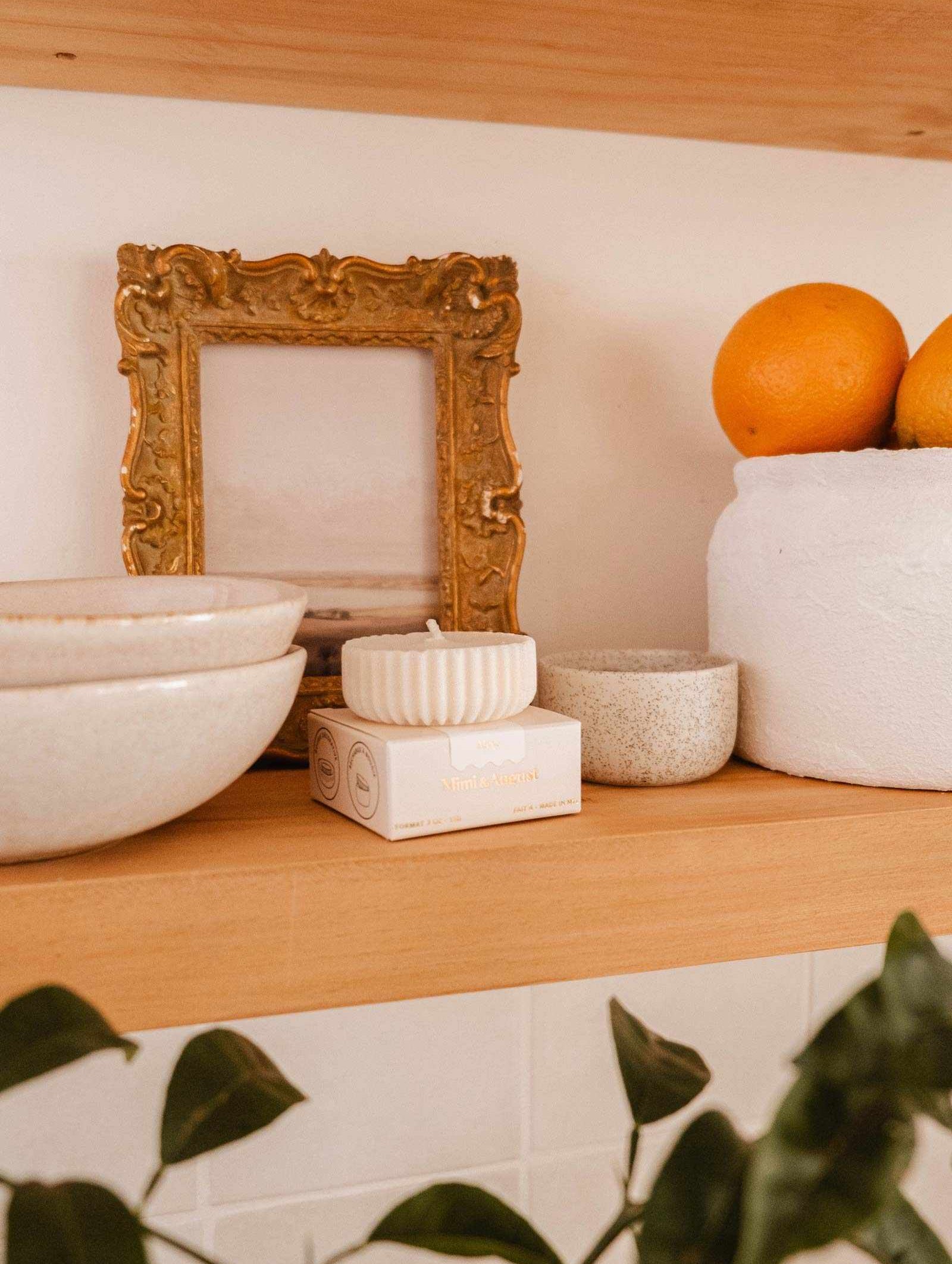 Wooden shelf with stacked ceramic bowls, a Mimi & August Candle Refill Alpes soy wax candle refill, a gold-framed picture, a white planter holding oranges, and a green plant partially visible in the foreground.