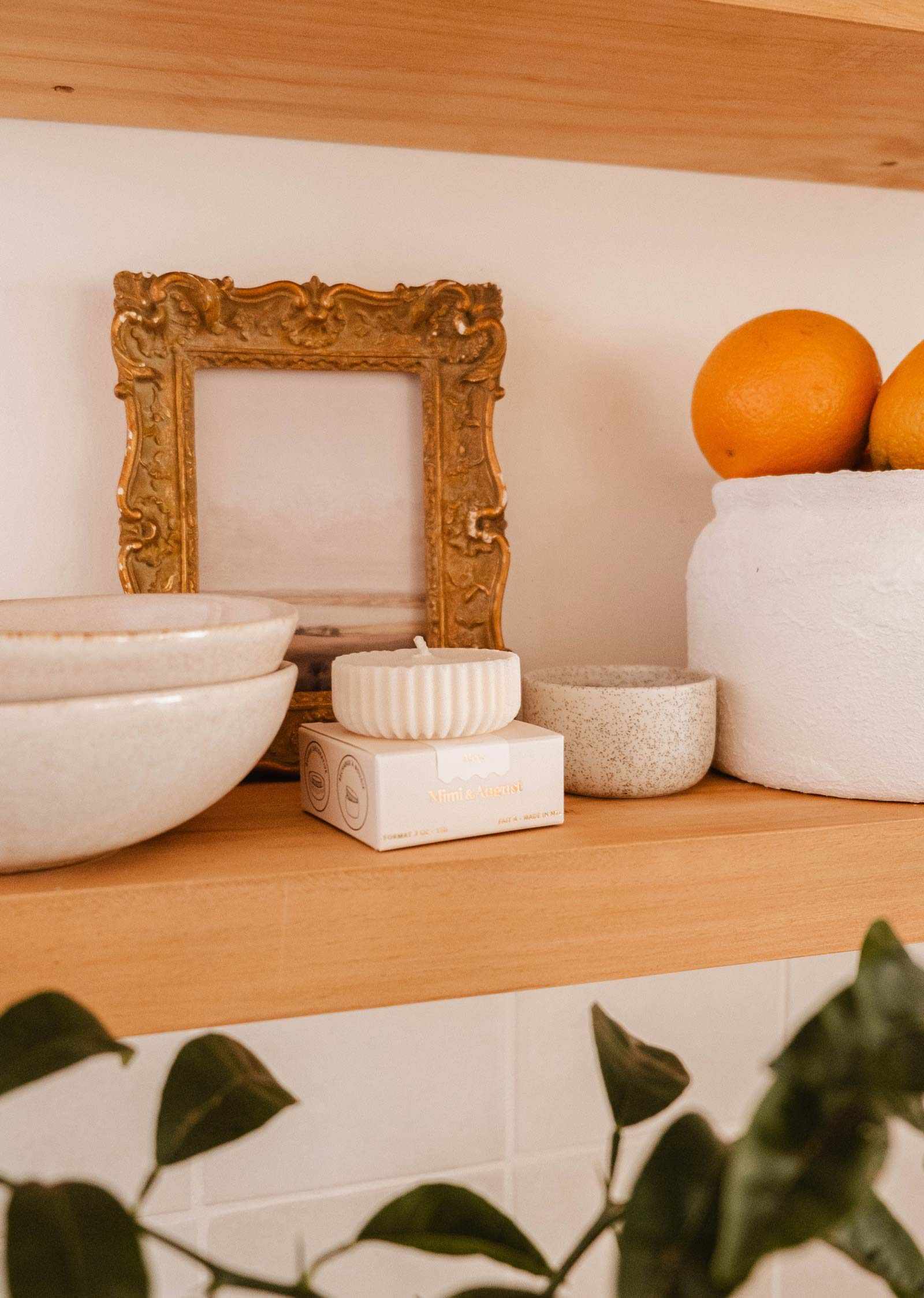 Wooden shelf with stacked ceramic bowls, a Mimi & August Candle Refill Alpes soy wax candle refill, a gold-framed picture, a white planter holding oranges, and a green plant partially visible in the foreground.
