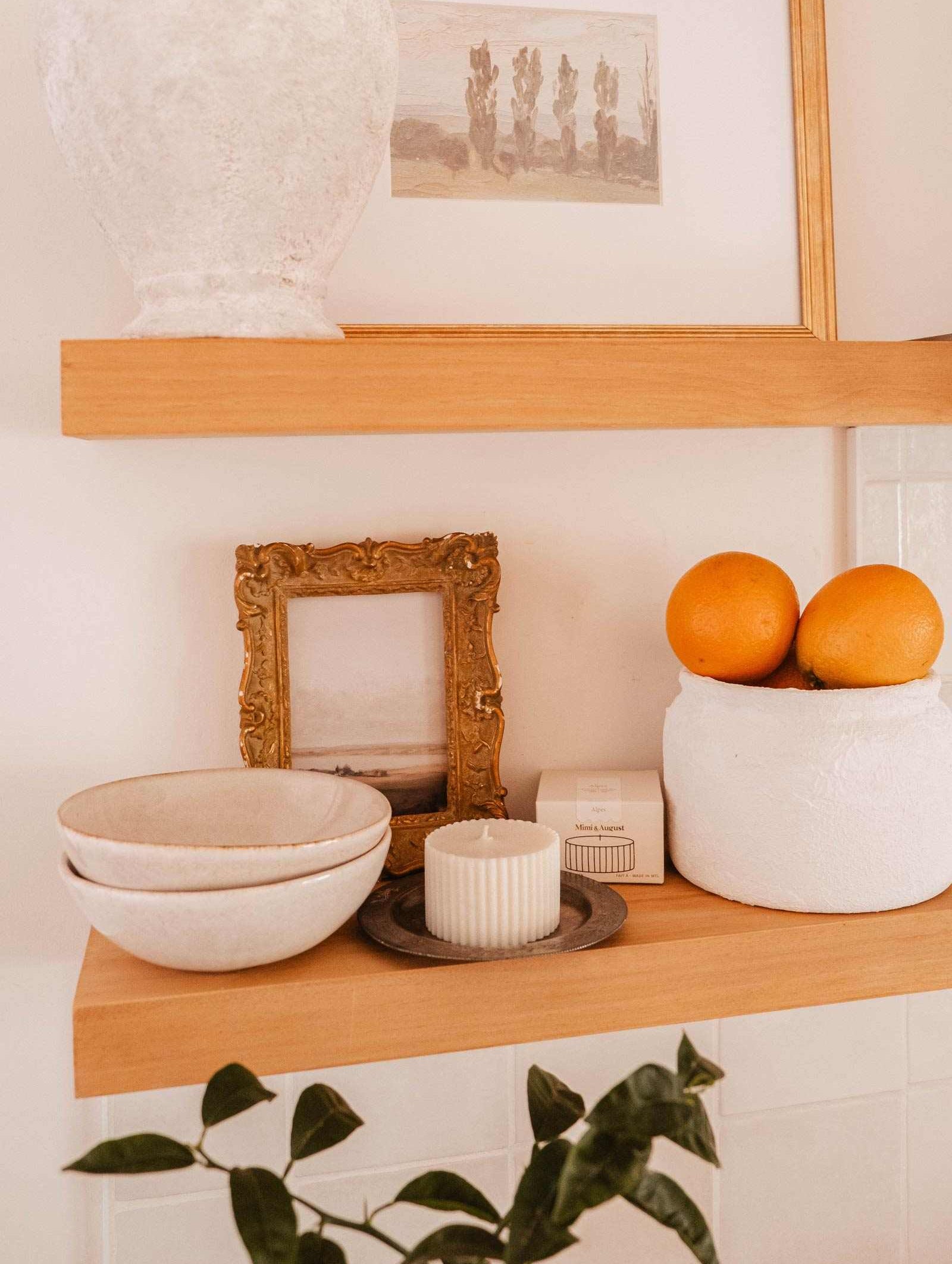 Wooden shelves display a white vase, framed pictures, a white container with two oranges, stacked bowls, the Mimi & August Candle Refill Alpes on a plate, plus a small box and a plant in the foreground.