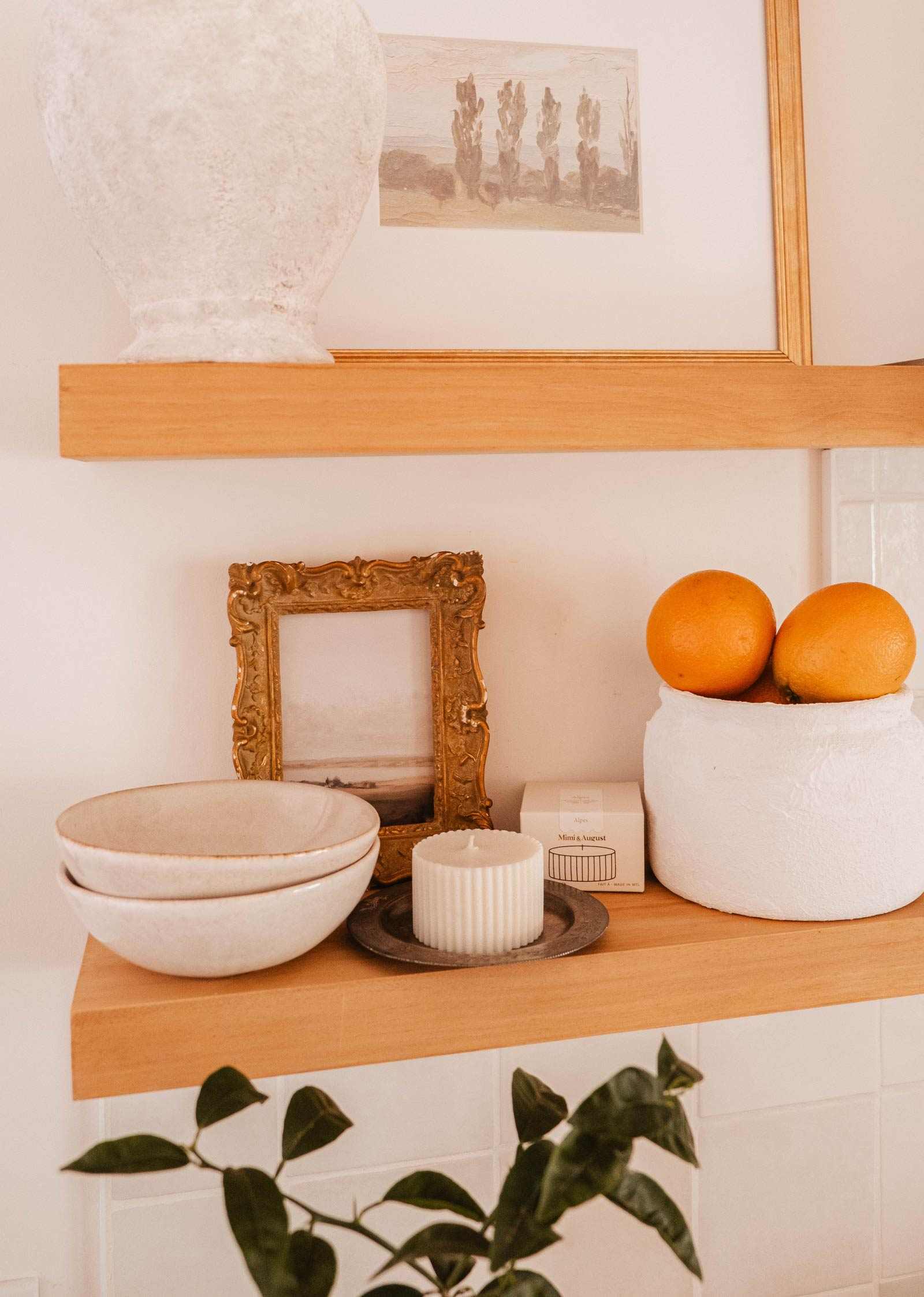 Wooden shelves display a white vase, framed pictures, a white container with two oranges, stacked bowls, the Mimi & August Candle Refill Alpes on a plate, plus a small box and a plant in the foreground.