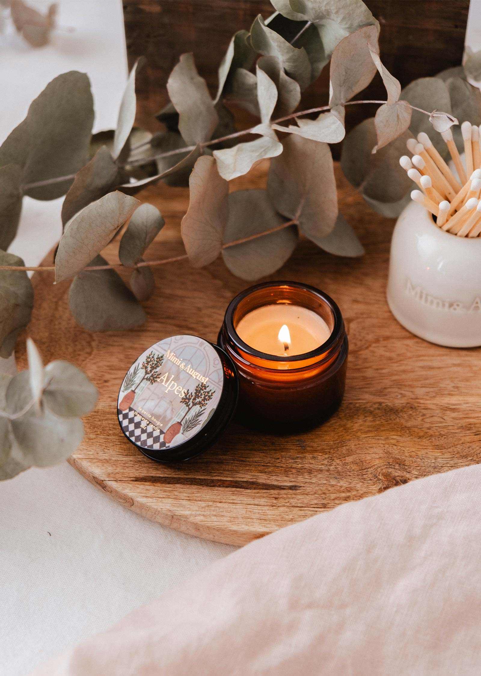 A lit Mimi & August Alpes - 2 oz reusable candle in amber glass rests on a wooden tray with dried eucalyptus leaves and a jar of matches, filling the air with an Alpine-fresh scent.