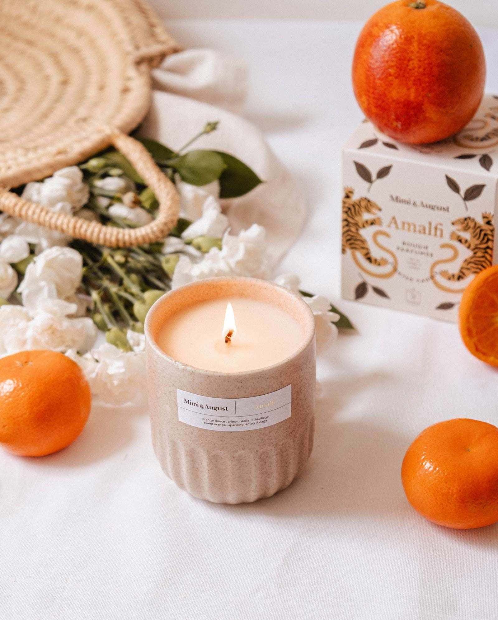 A lit Mimi & August Amalfi - Reusable Candle in a beige container rests on a white surface, surrounded by oranges, white flowers, tropical leaves, a woven bag, and an “Amalfi” box—evoking the spirit of seaside holidays.