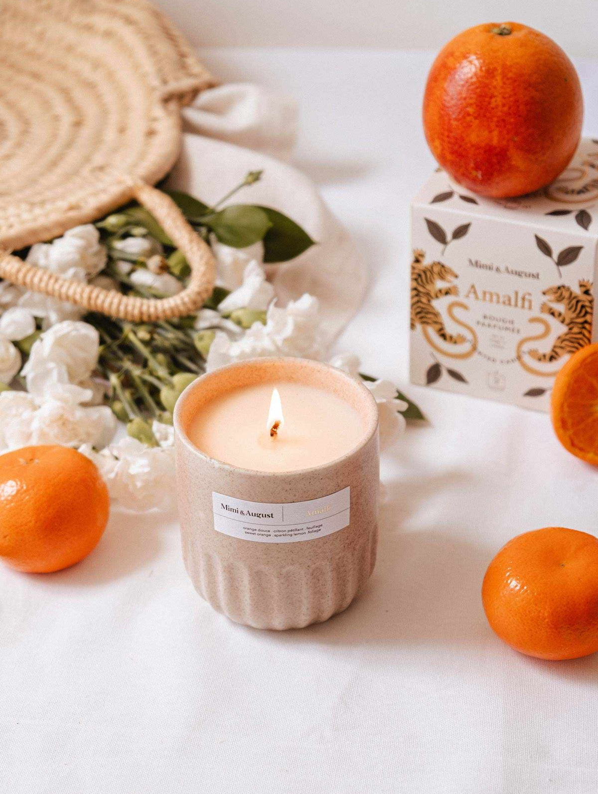 A lit Mimi & August Amalfi - Reusable Candle in a beige container rests on a white surface, surrounded by oranges, white flowers, tropical leaves, a woven bag, and an “Amalfi” box—evoking the spirit of seaside holidays.