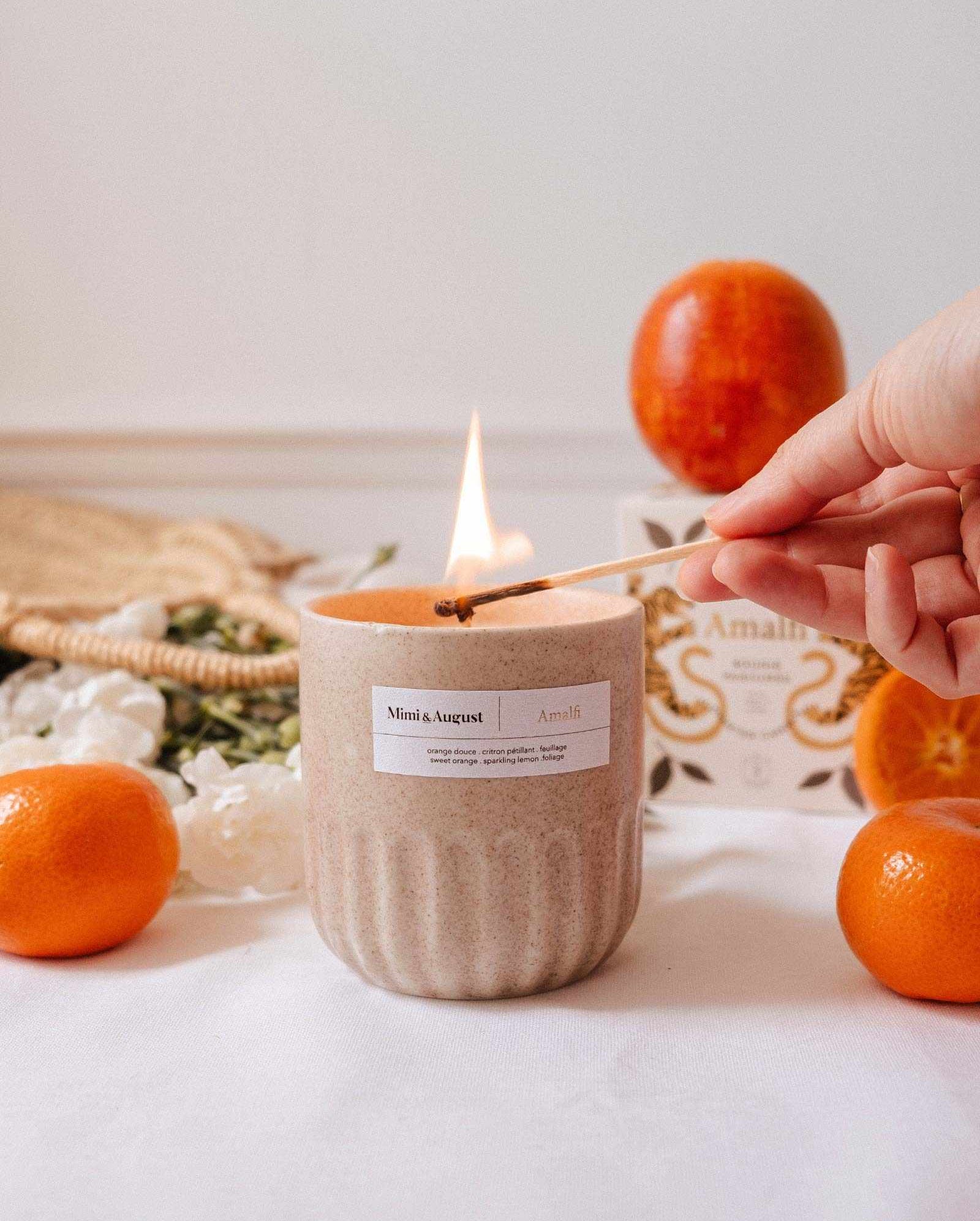 A hand lights the Amalfi - Reusable Candle by Mimi & August in a beige jar on a white surface, with oranges, a box, and tropical foliage nearby.