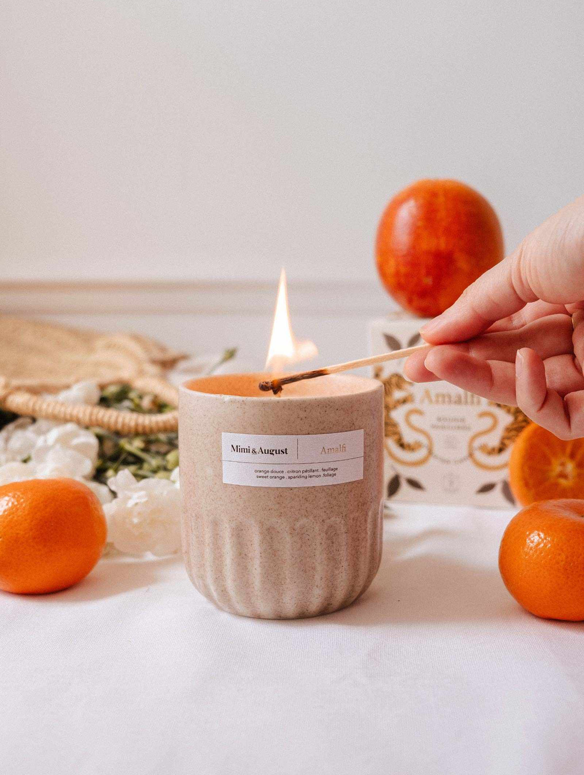 A hand lights the Amalfi - Reusable Candle by Mimi & August in a beige jar on a white surface, with oranges, a box, and tropical foliage nearby.