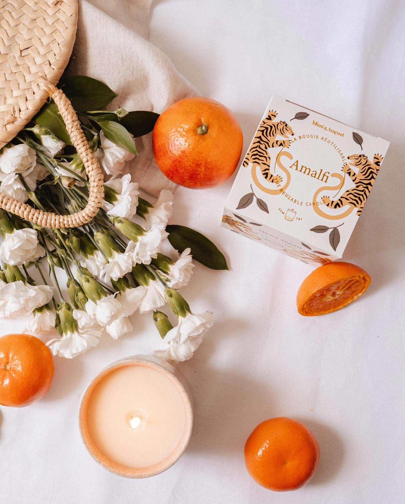 A woven bag with white flowers, oranges, and a lit Mimi & August Amalfi - Reusable Candle evokes memories of seaside holidays, all beautifully arranged on a white surface.