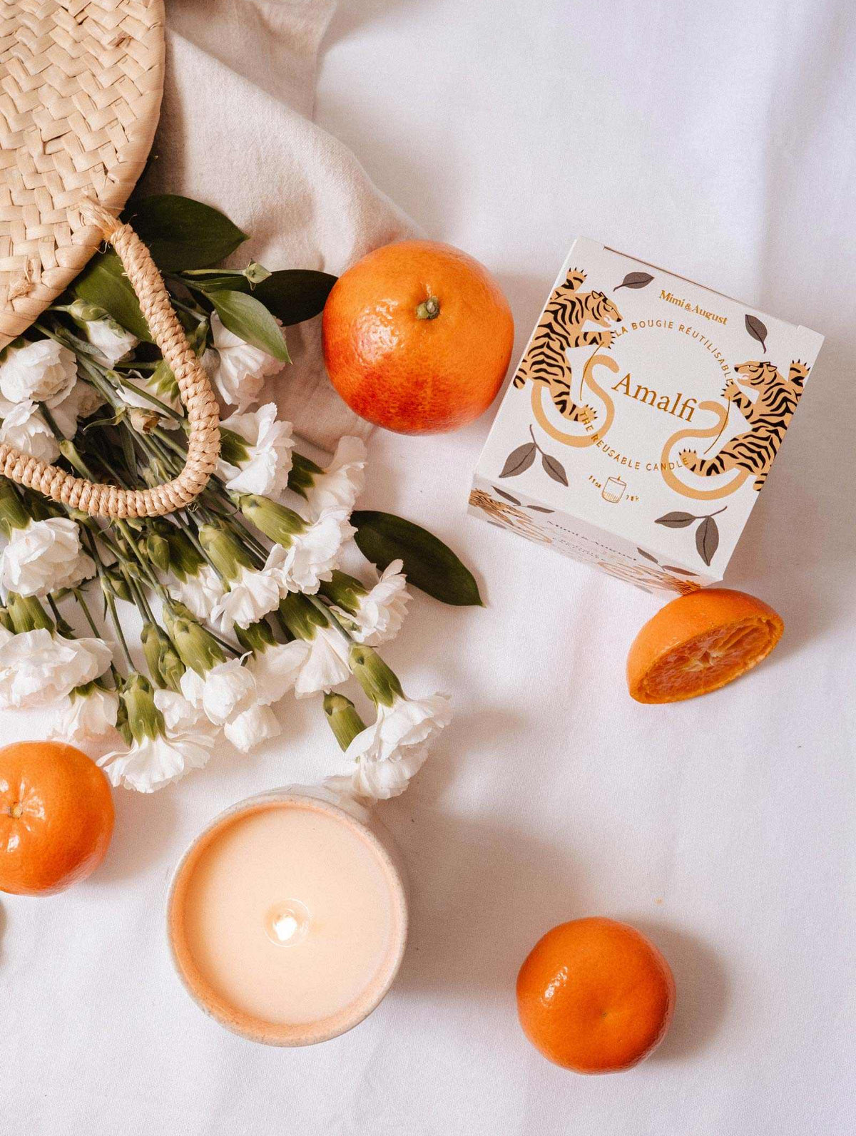 A woven bag with white flowers, oranges, and a lit Mimi & August Amalfi - Reusable Candle evokes memories of seaside holidays, all beautifully arranged on a white surface.
