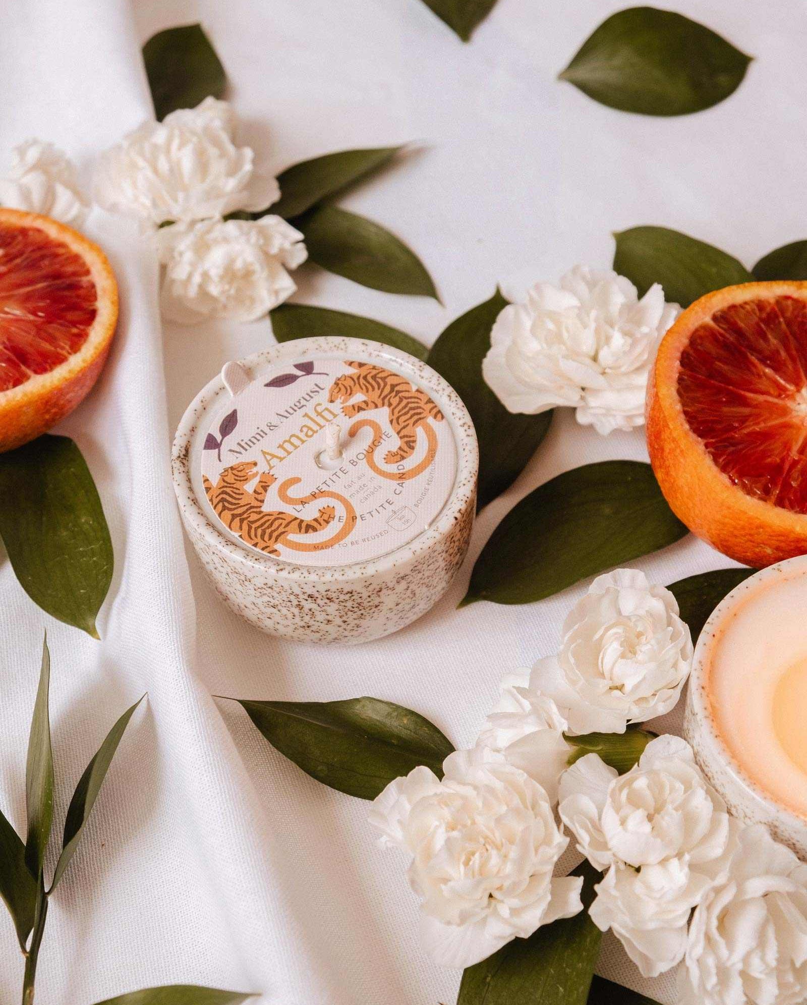 A round "Amalfi - Reusable Candle" by Mimi & August rests amid white flowers, tropical leaves, and blood orange slices on white fabric, capturing the essence of seaside holidays.