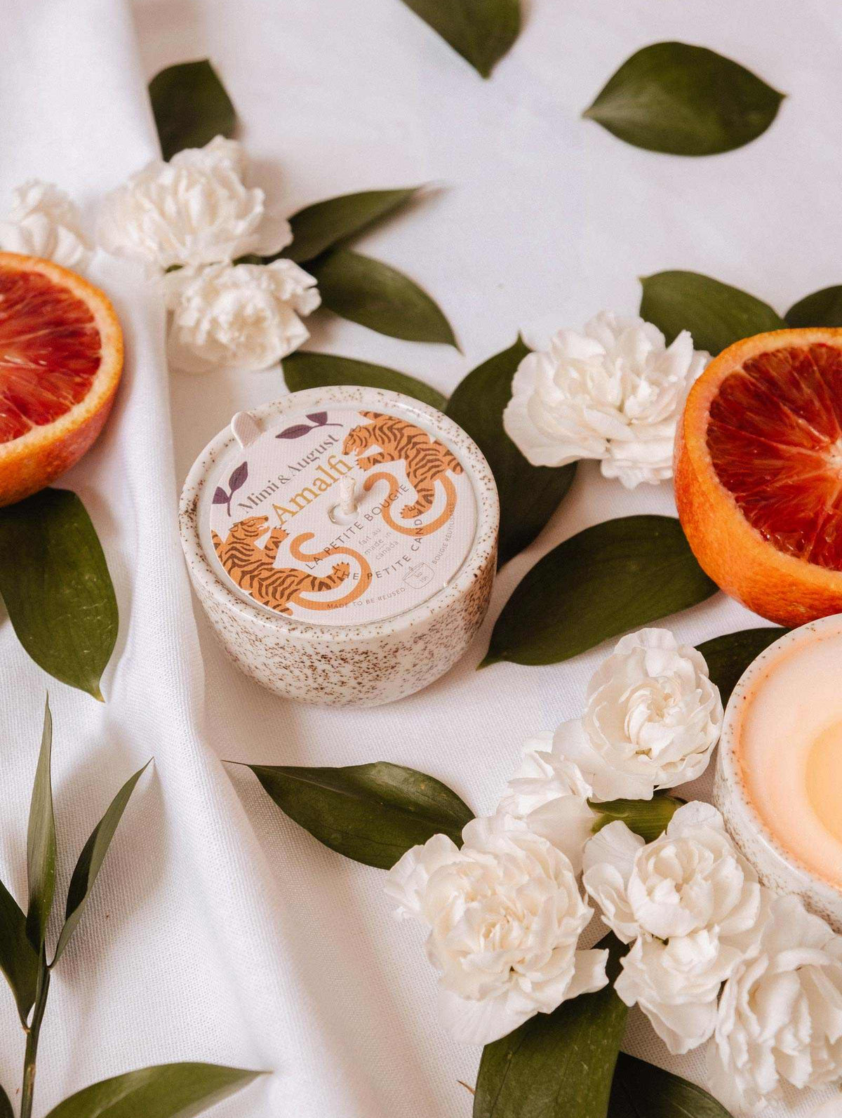 A round "Amalfi - Reusable Candle" by Mimi & August rests amid white flowers, tropical leaves, and blood orange slices on white fabric, capturing the essence of seaside holidays.