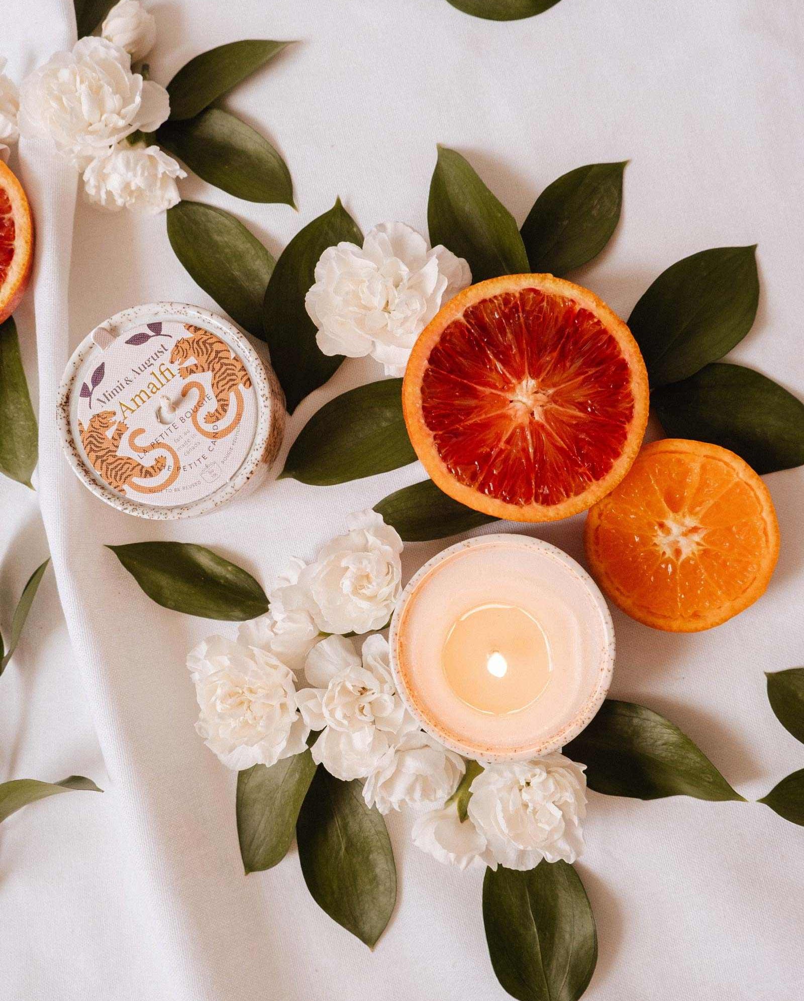 A lit Mimi & August Amalfi - Reusable Candle, sliced blood orange and orange, a round jar, white flowers, and lush tropical foliage are arranged on a white surface.