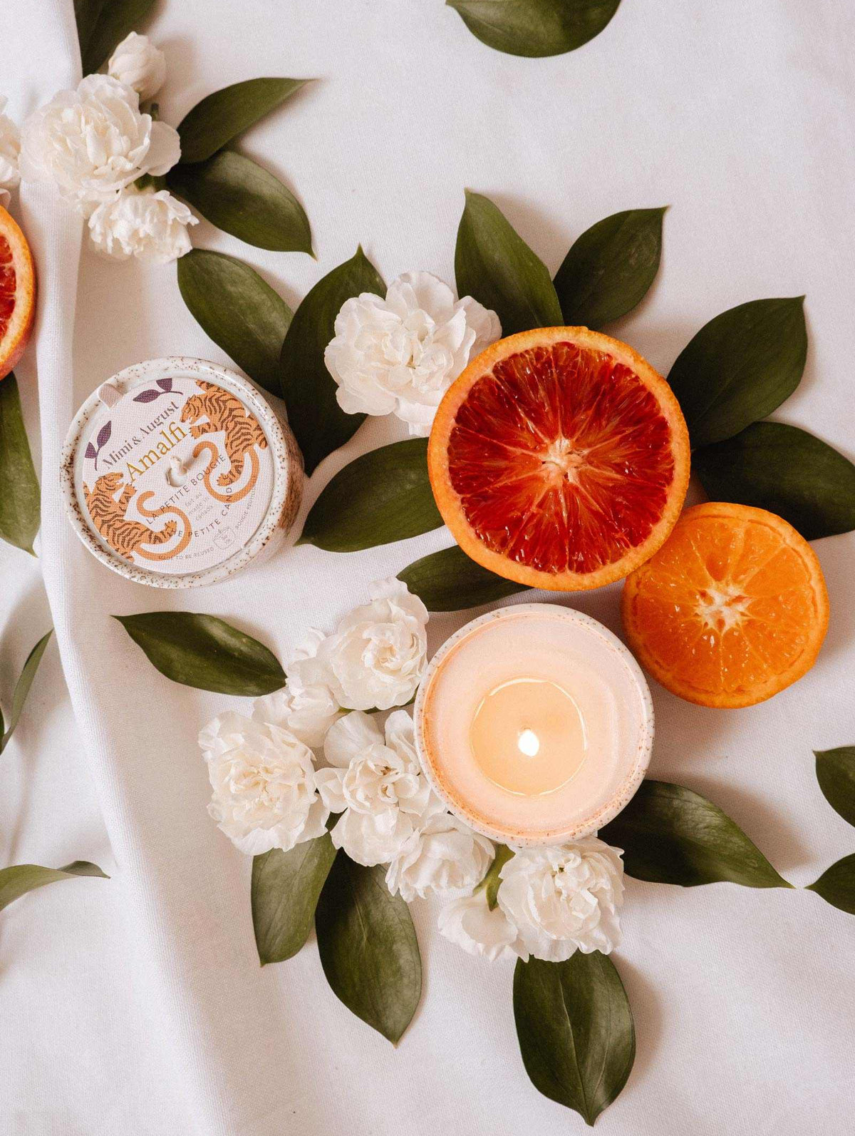 A lit Mimi & August Amalfi - Reusable Candle, sliced blood orange and orange, a round jar, white flowers, and lush tropical foliage are arranged on a white surface.