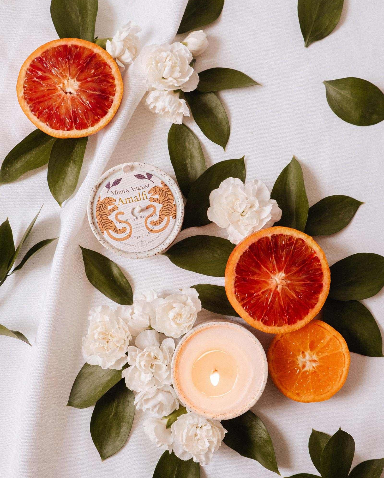 Sliced blood oranges, white flowers, tropical leaves, and the Mimi & August Amalfi - Reusable Candle are beautifully arranged on white fabric, evoking seaside holiday memories.