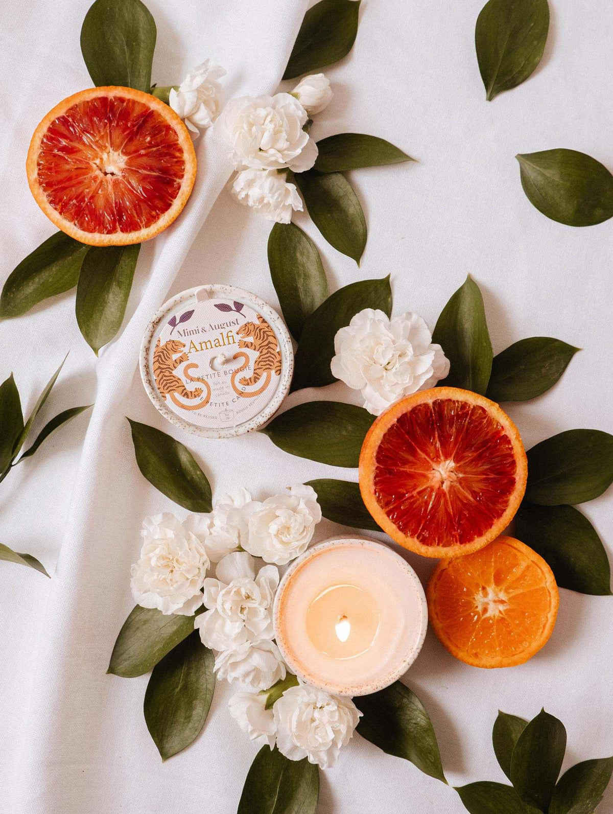 Sliced blood oranges, white flowers, tropical leaves, and the Mimi & August Amalfi - Reusable Candle are beautifully arranged on white fabric, evoking seaside holiday memories.