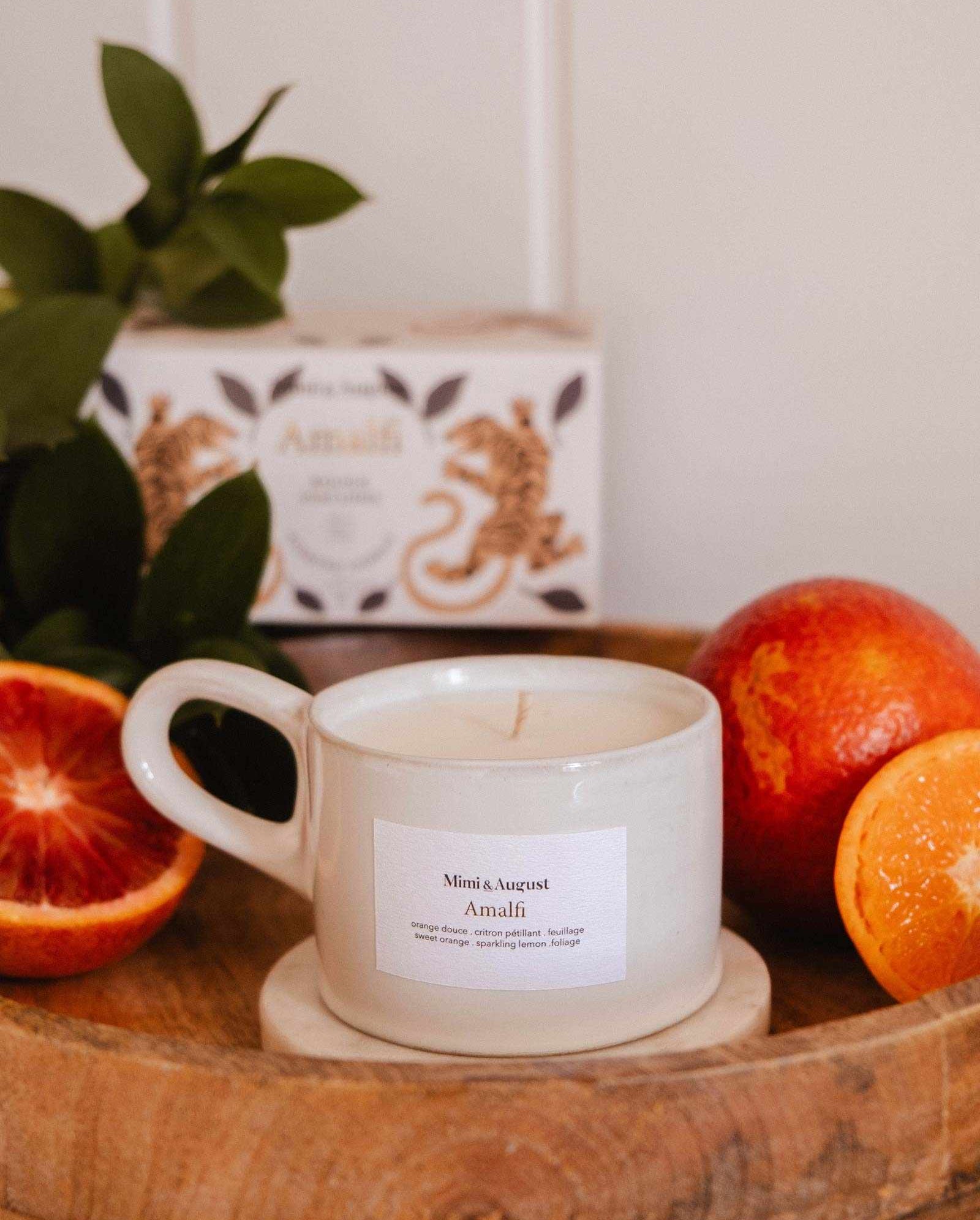 A white Mimi & August Amalfi - Reusable Candle in a cup rests on a wooden tray with blood oranges, tropical leaves, and a decorative box, capturing the seaside holiday charm.