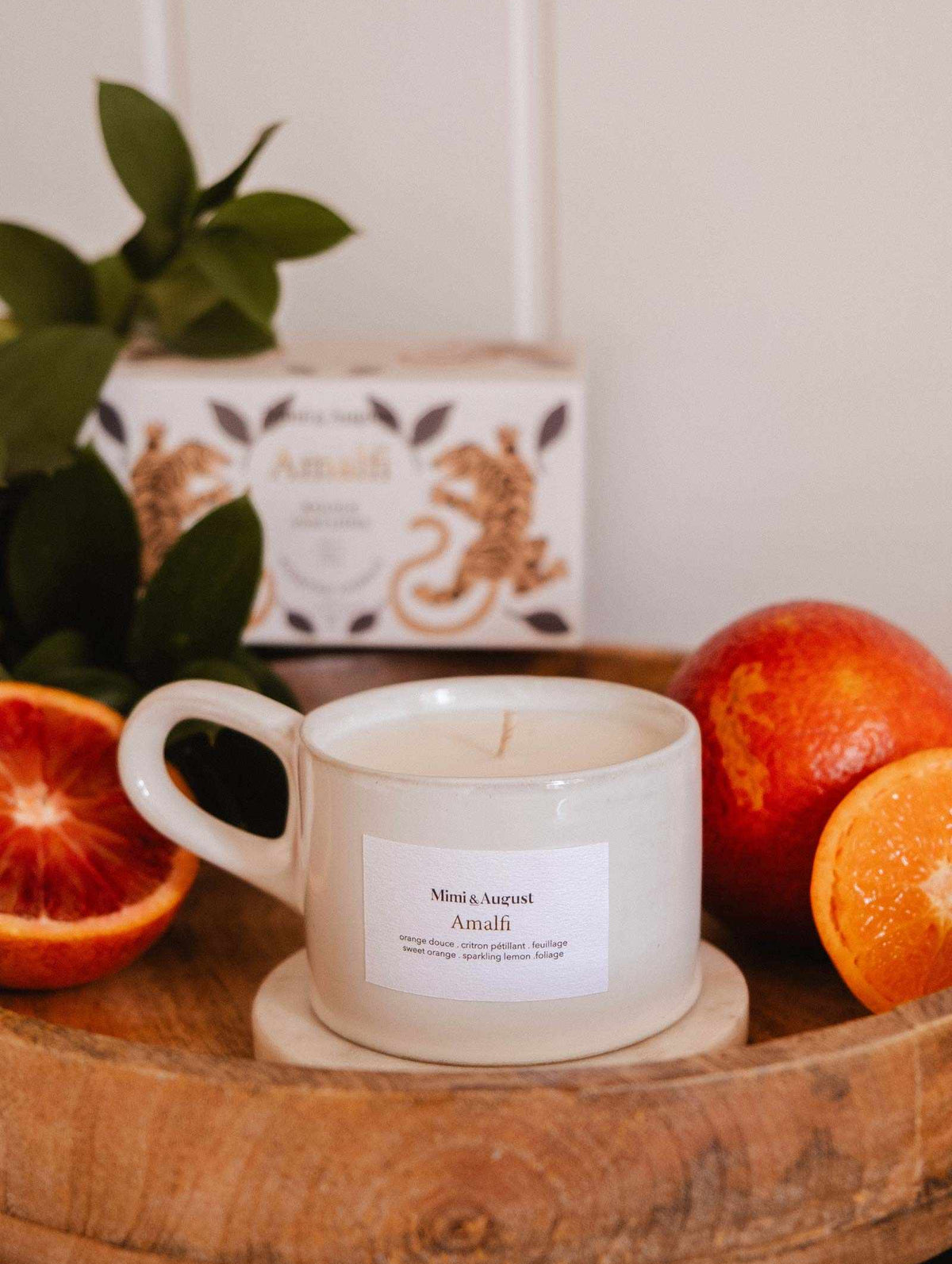 A white Mimi & August Amalfi - Reusable Candle in a cup rests on a wooden tray with blood oranges, tropical leaves, and a decorative box, capturing the seaside holiday charm.