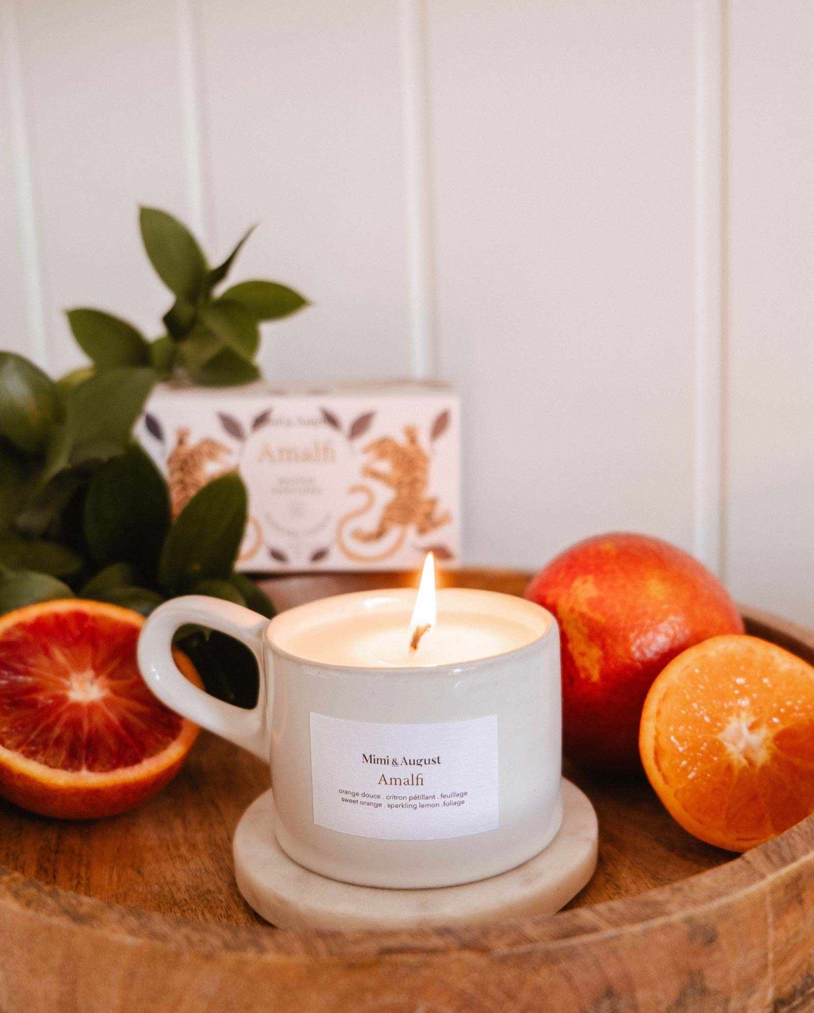 A lit Amalfi - Reusable Candle by Mimi & August, in a white cup on a wooden tray with a marble coaster, is surrounded by blood oranges and tropical foliage, evoking seaside holidays. An "Amali" box appears in the background.