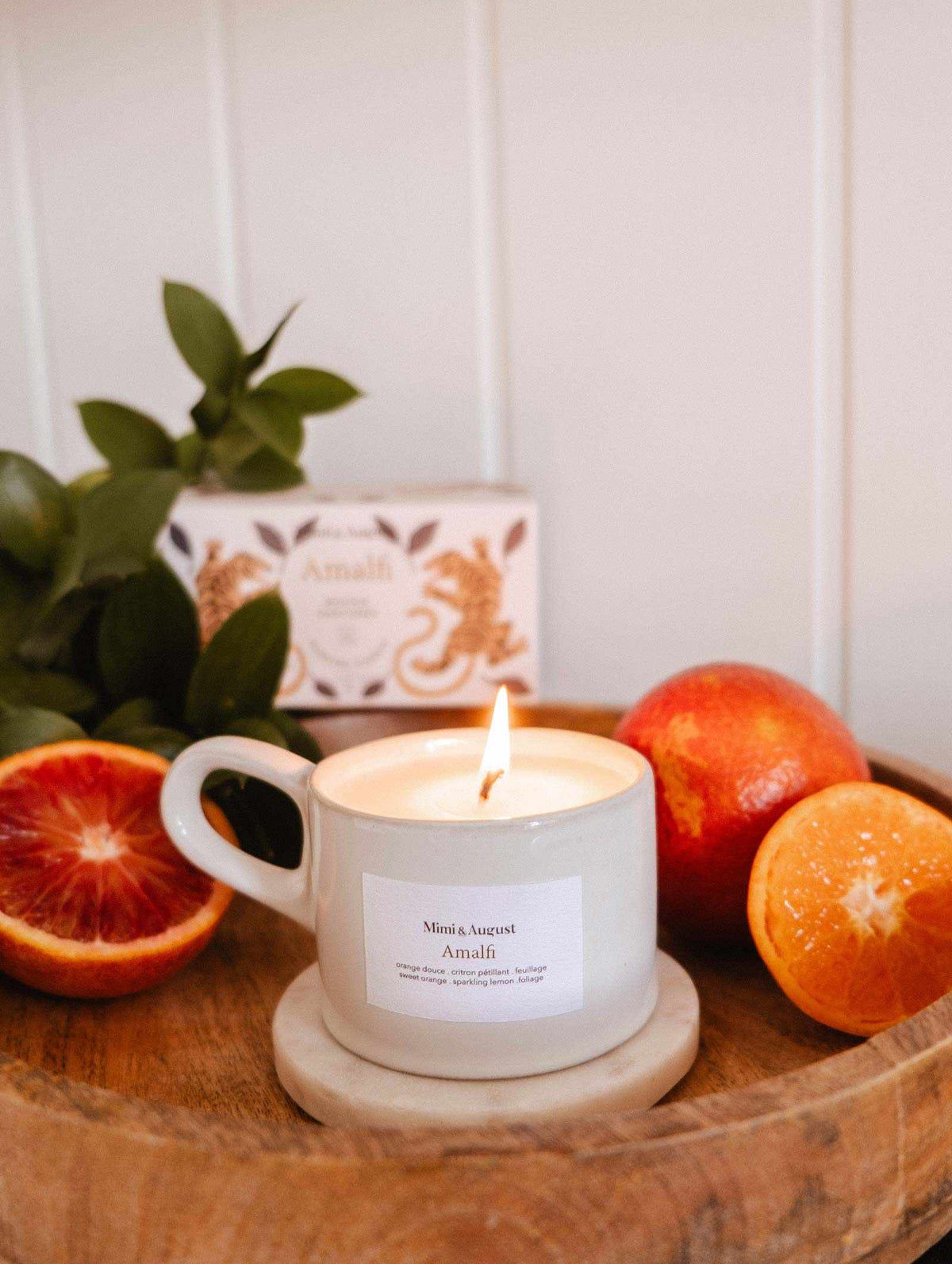 A lit Amalfi - Reusable Candle by Mimi & August, in a white cup on a wooden tray with a marble coaster, is surrounded by blood oranges and tropical foliage, evoking seaside holidays. An "Amali" box appears in the background.