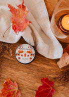 A tin labeled "Ambre - Reusable Candle" by Mimi & August rests on a wooden surface surrounded by autumn leaves, a lit candle radiating fall coziness, and a white cloth nearby, creating a tranquil atmosphere.