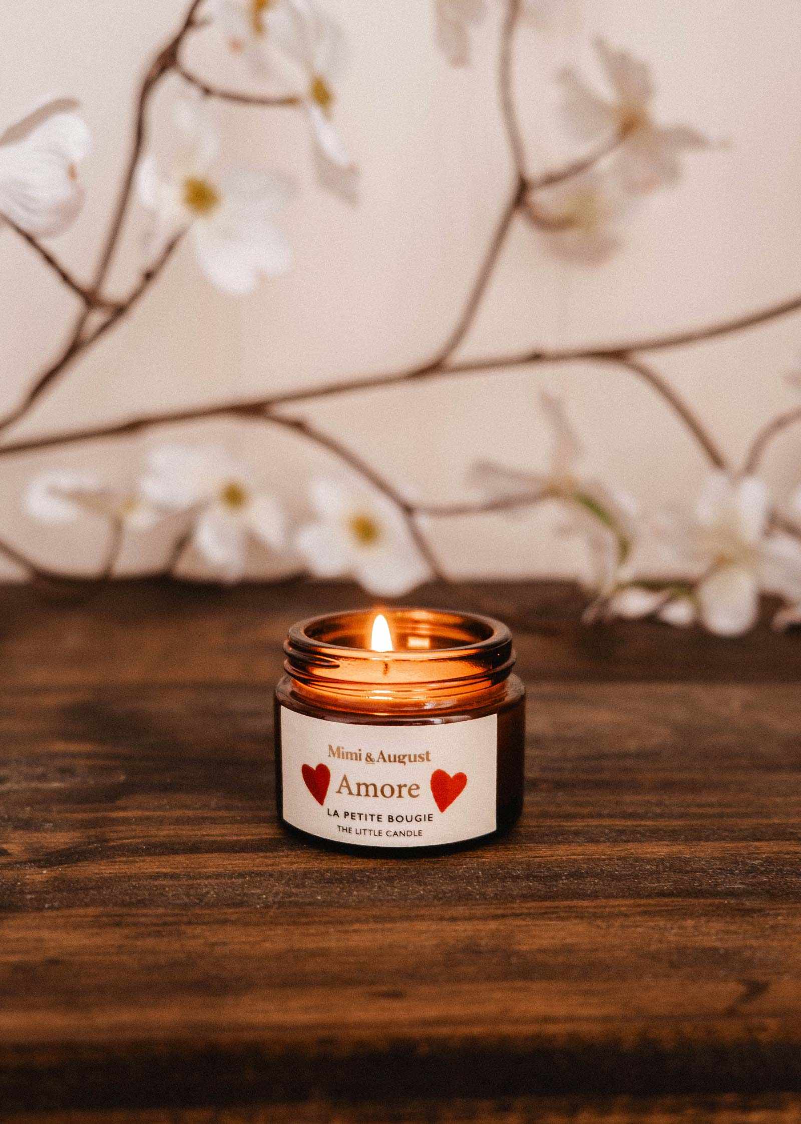 The Amore – 2 oz Soy Candle by Mimi & August sits on a wooden surface, surrounded by white flowers and branches in the background.