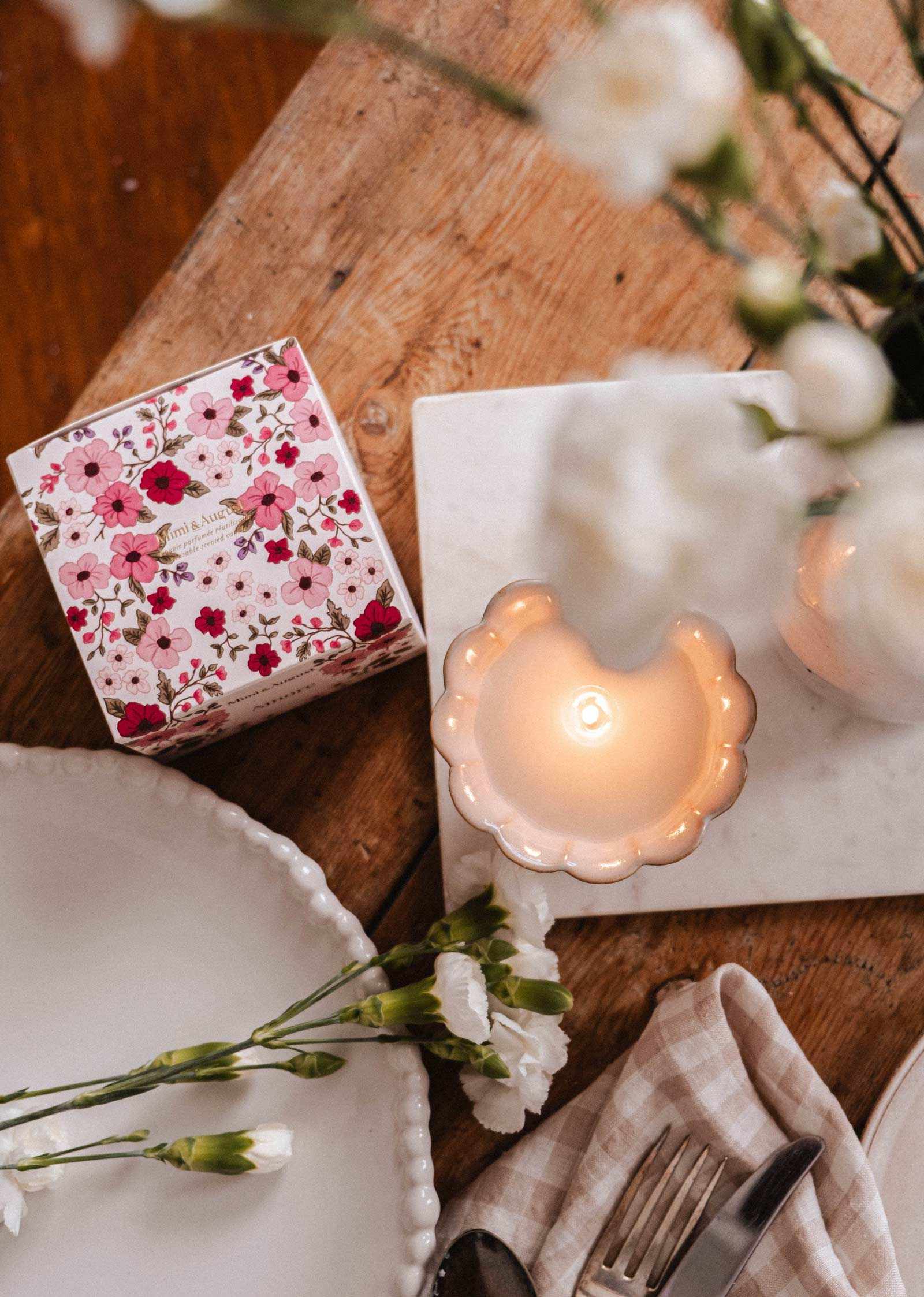 The Amore Reusable Candle by Mimi & August, a floral-patterned box, white flowers, plates, and cutlery are displayed on a wooden table with a marble tray.