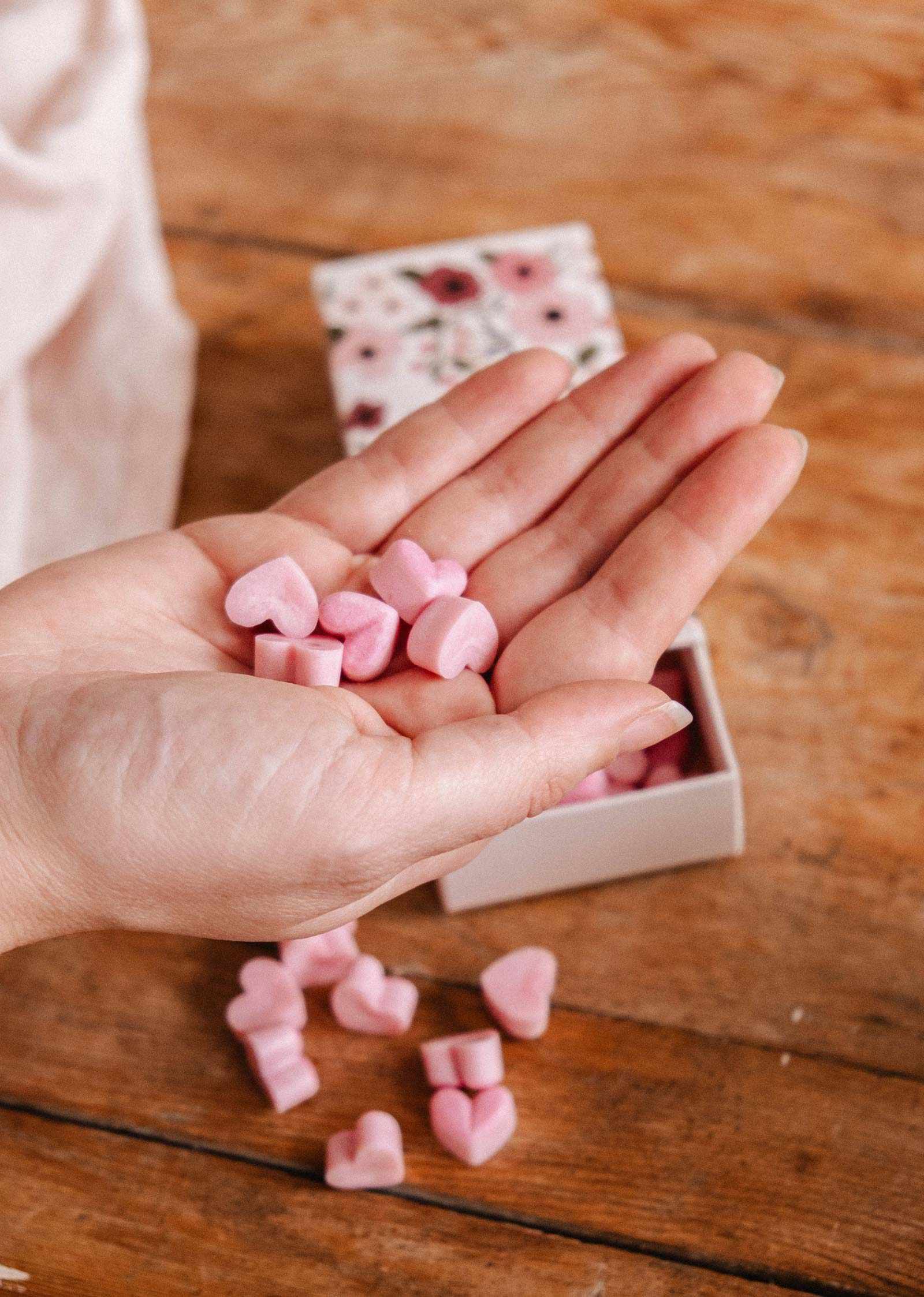 A hand holds several small pink heart-shaped Amore Scented Wax Melts by Mimi & August above a wooden surface, with more melts and a floral Mimi & August box in the background.