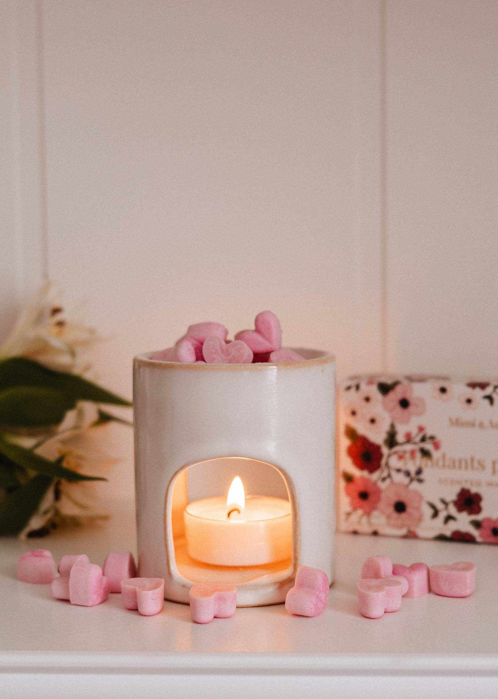 A white ceramic candle holder with a lit tealight, filled with Mimi & August’s Amore Scented Wax Melts in pink heart shapes, sits on a white surface with scattered melts and a floral box in the background, creating a romantic aroma.