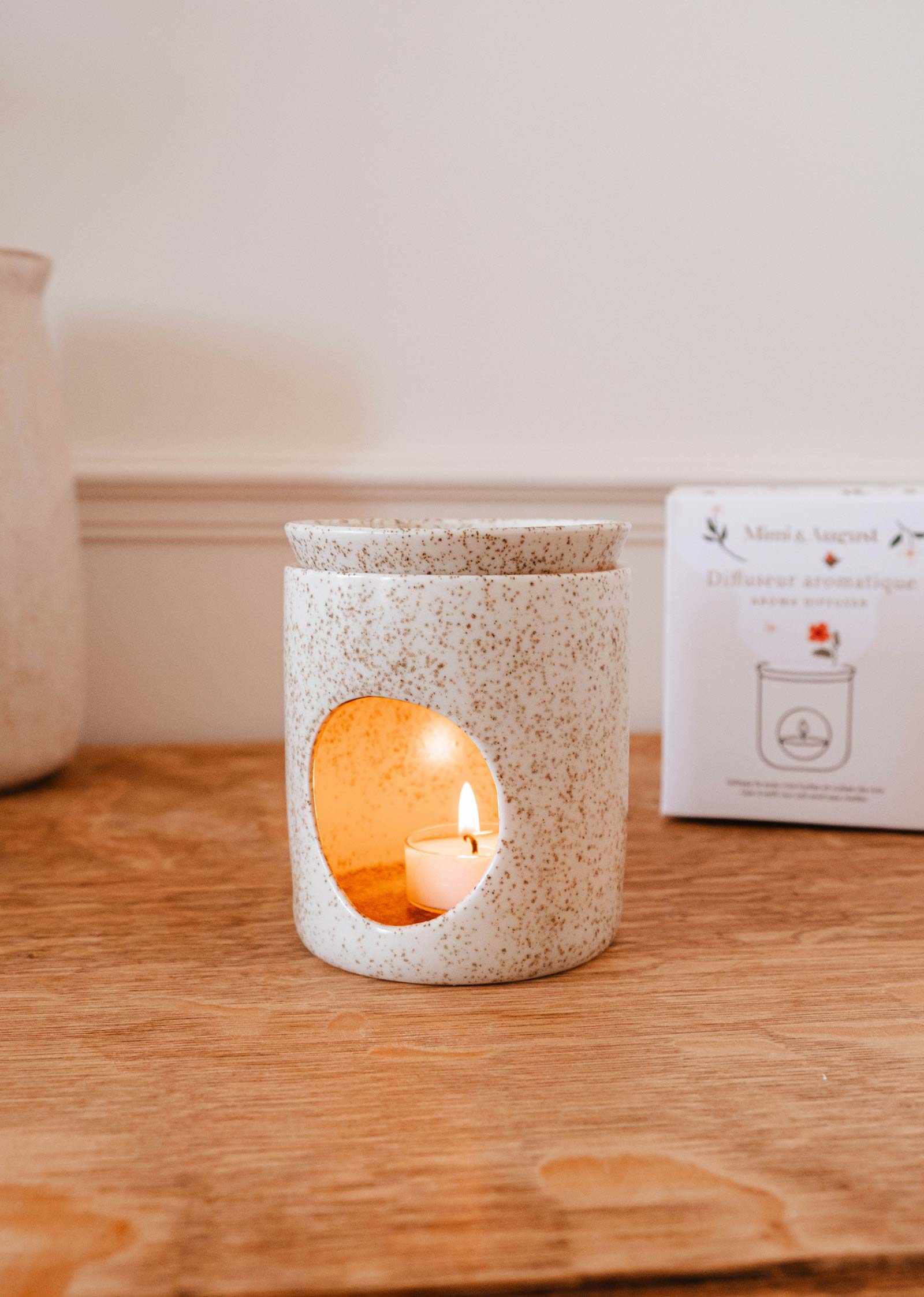 An Aromatic Ceramic Diffuser by Mimi & August, featuring a white speckled design and a candle inside, sits on a wooden surface with the product box in the background.