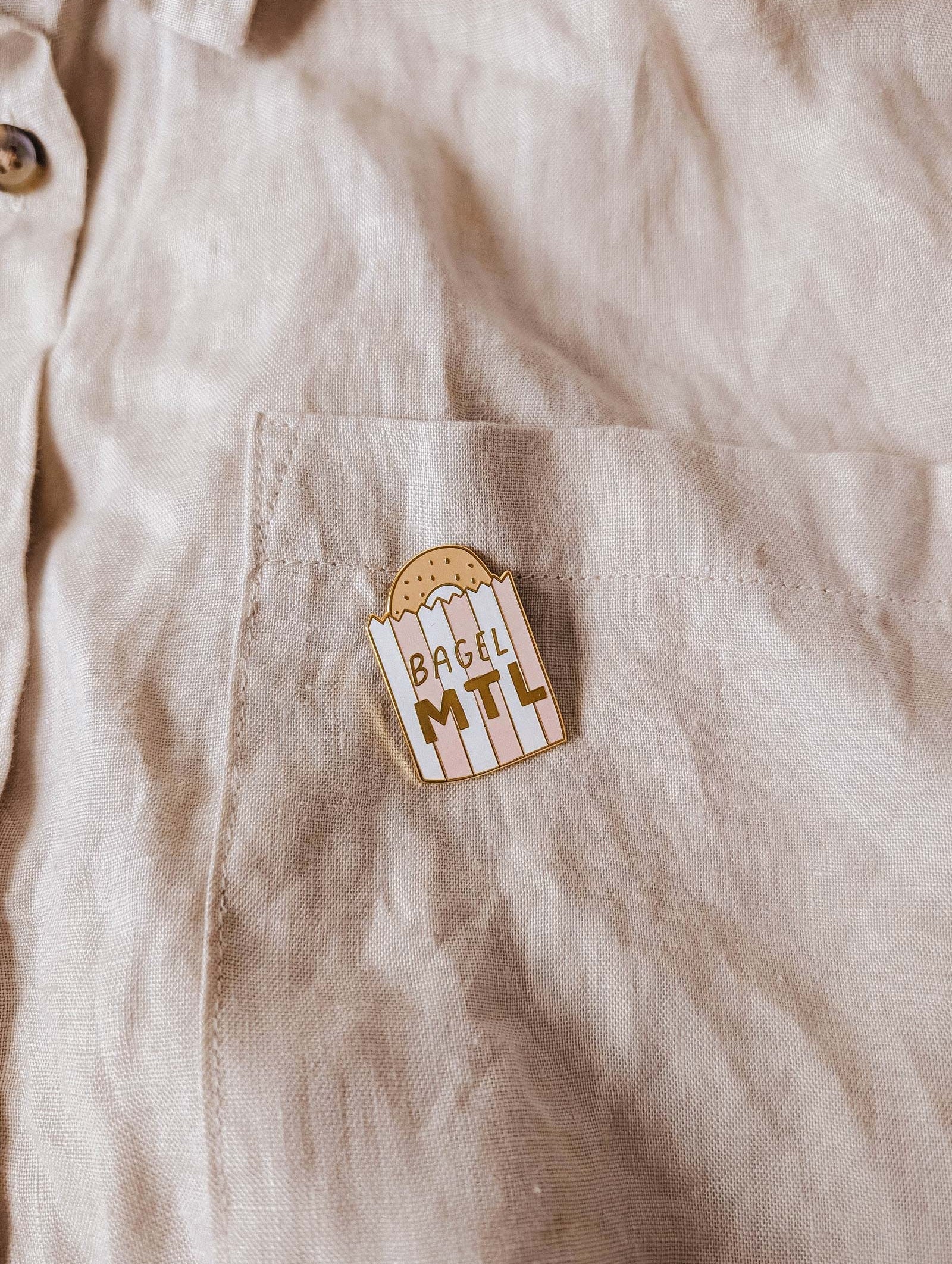 The beige shirt pocket displays the Mimi & August Bagel MTL Enamel Pin, featuring a bagel graphic and "BAGEL MTL" text—an ideal accessory for Montréal bagel fans and food lovers.