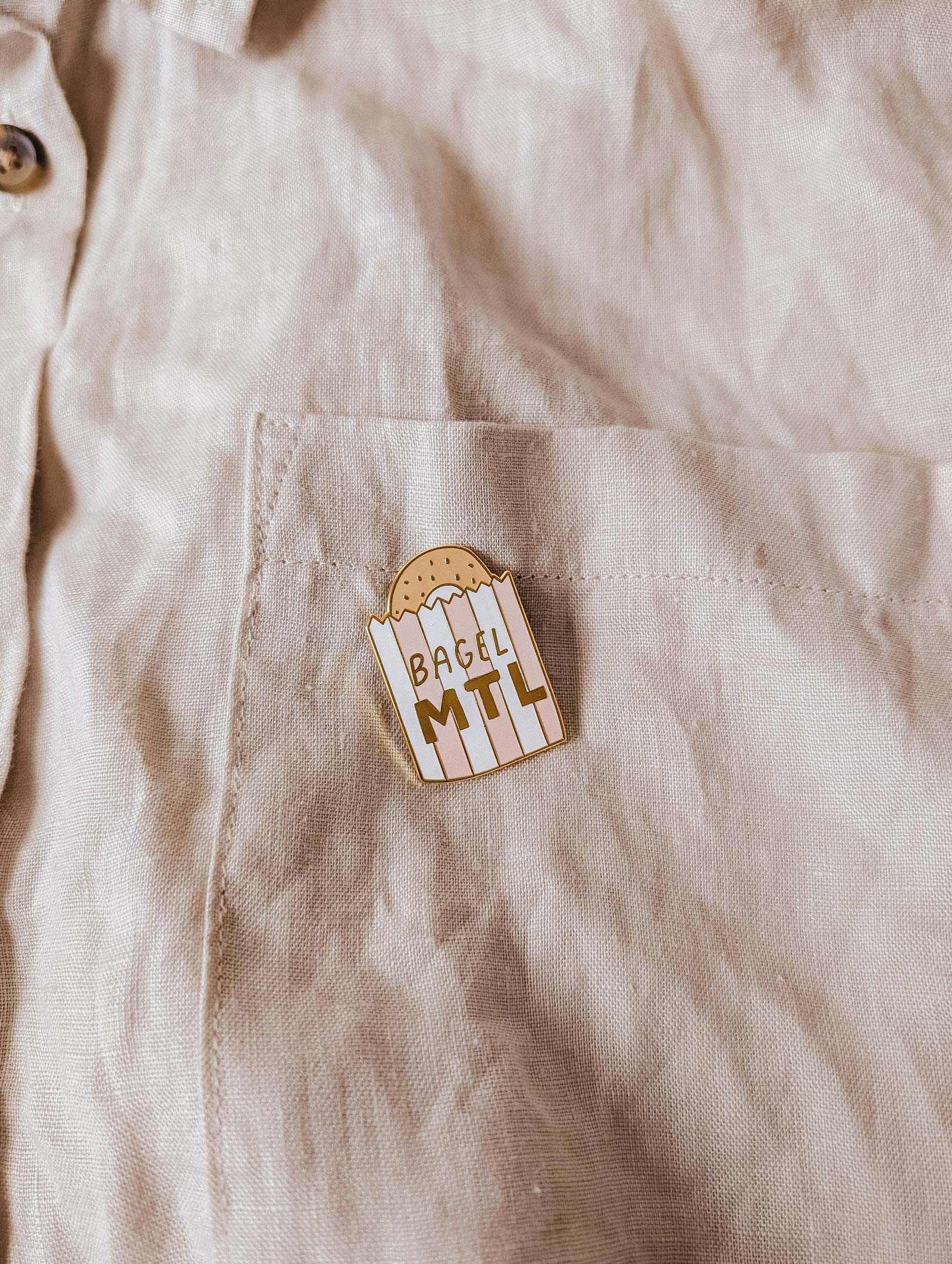 The beige shirt pocket displays the Mimi & August Bagel MTL Enamel Pin, featuring a bagel graphic and "BAGEL MTL" text—an ideal accessory for Montréal bagel fans and food lovers.