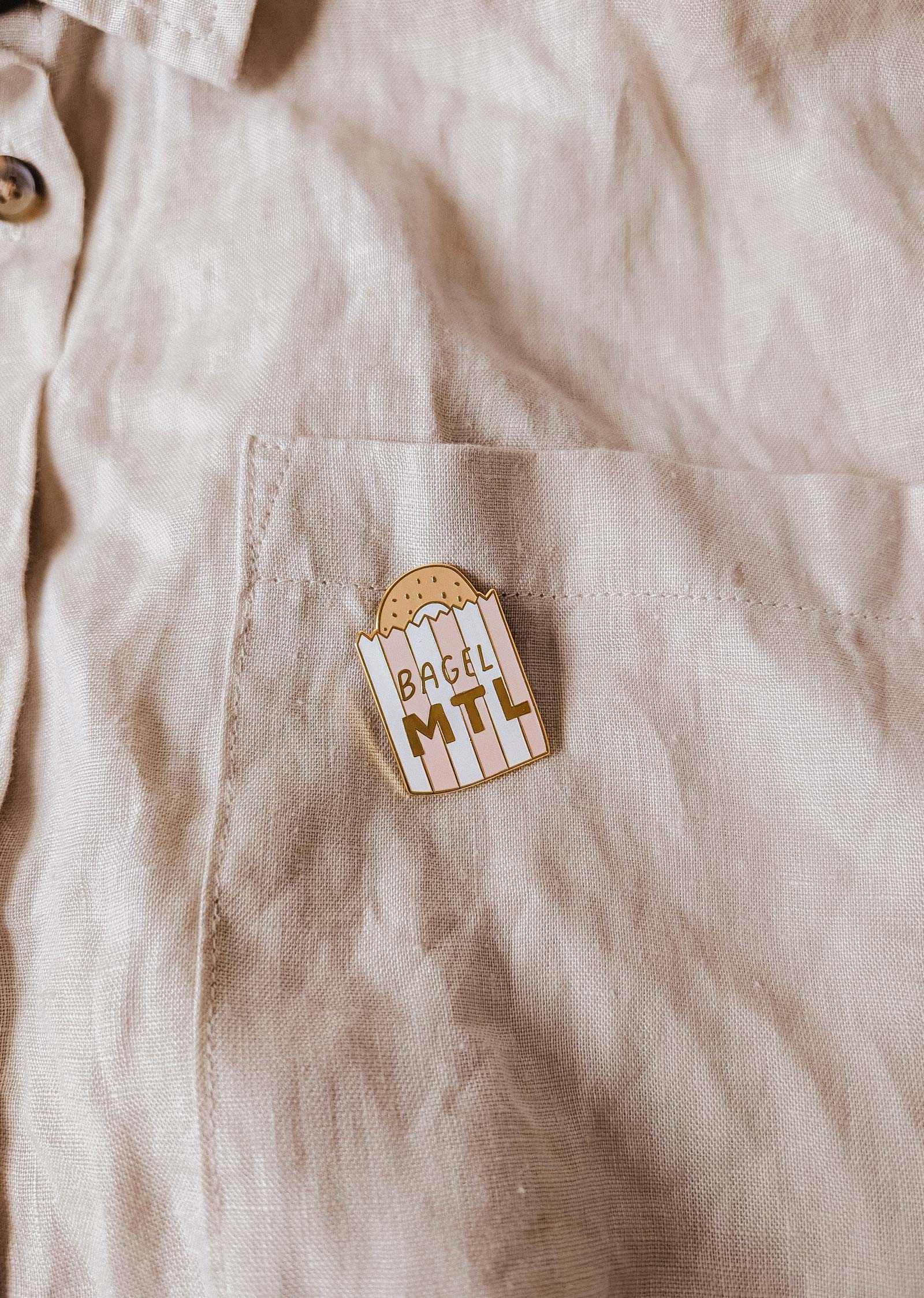 The beige shirt pocket displays the Mimi & August Bagel MTL Enamel Pin, featuring a bagel graphic and "BAGEL MTL" text—an ideal accessory for Montréal bagel fans and food lovers.