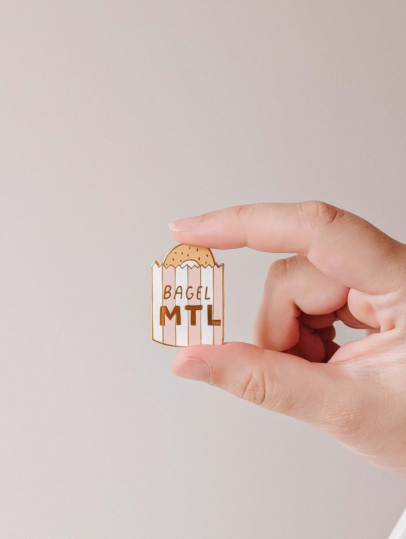 A hand holds a Bagel MTL Enamel Pin by Mimi & August, shaped like a Montréal bagel bag with "BAGEL MTL" on it—an ideal accessory for foodies and bagel lovers.