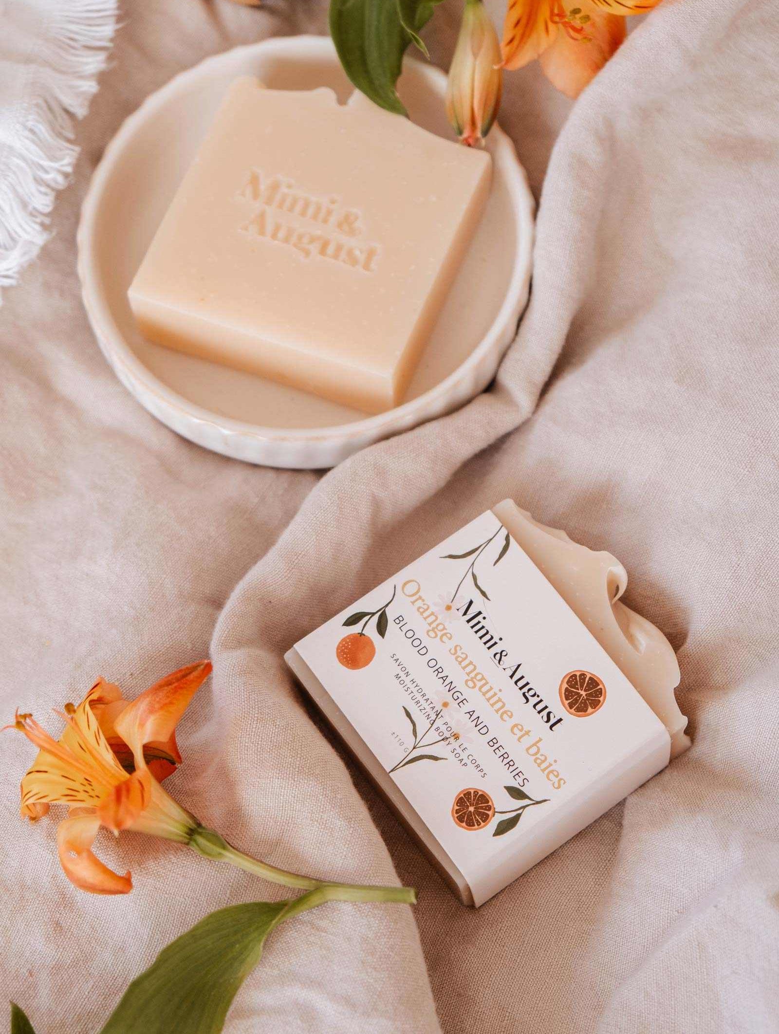 Aesthetic arrangement of hydrating Mimi & August Bar Soap - Blood Orange & Berries with floral decorations on a soft fabric surface.