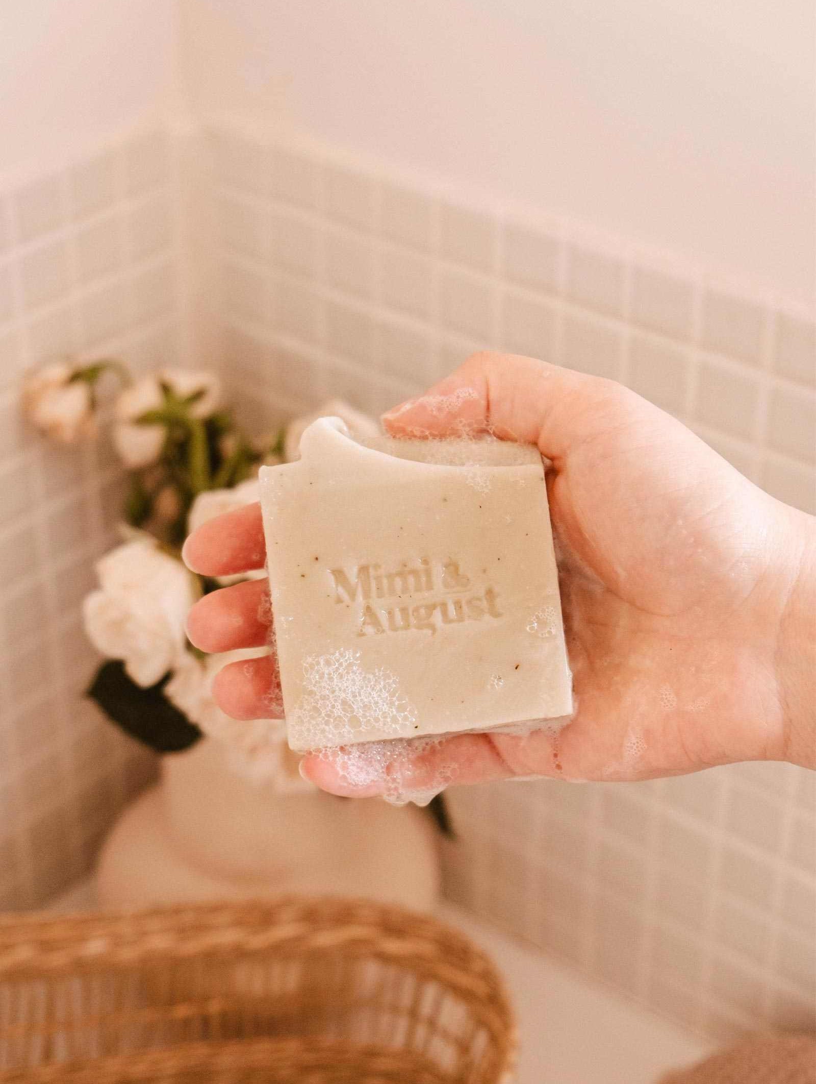 A person holds a bar of Mimi & August Eucalyptus Bar Soap over a bathtub with a floral arrangement and a woven basket in the background.