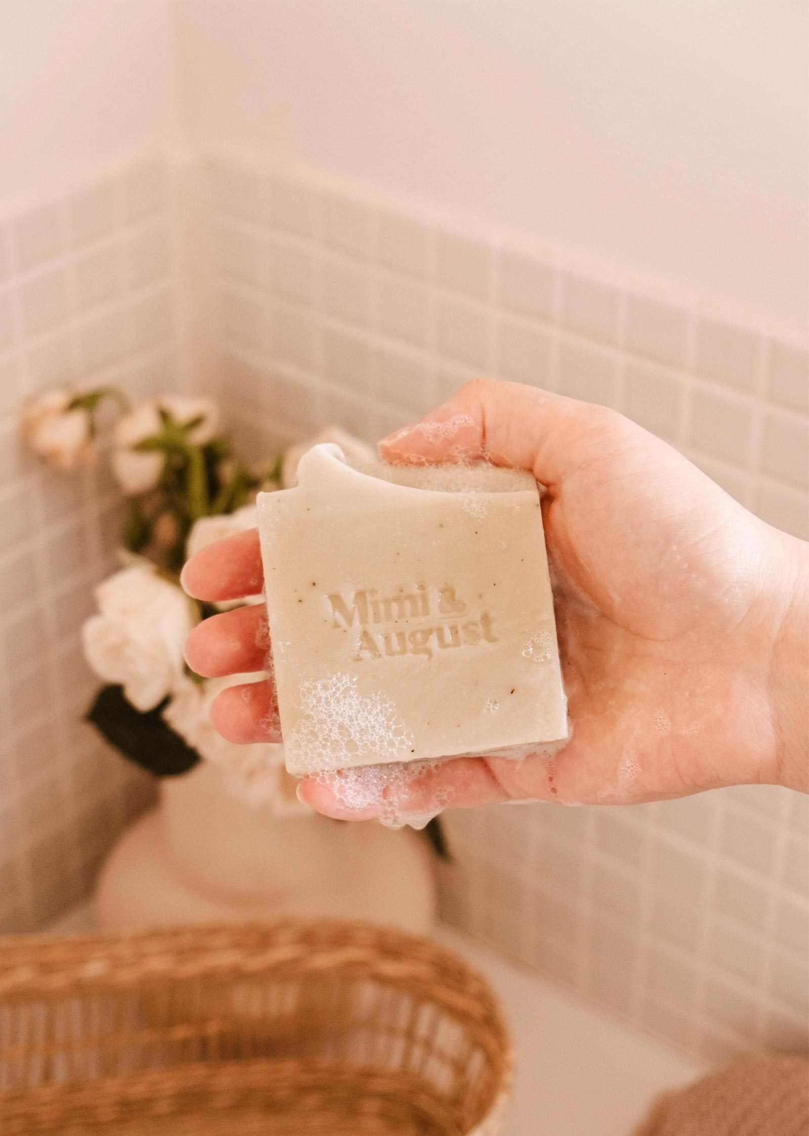 A person holds a bar of Mimi & August Eucalyptus Bar Soap over a bathtub with a floral arrangement and a woven basket in the background.