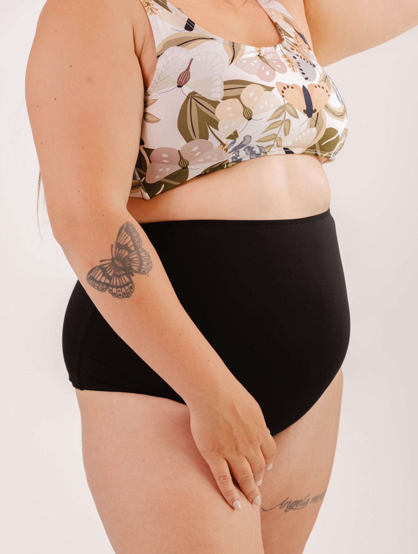 A person wearing a floral bikini top and Bermudes Black extra high waisted bikini bottom by Mimi & August shows off a tattoo of a butterfly on their upper arm, showcasing a flattering silhouette.