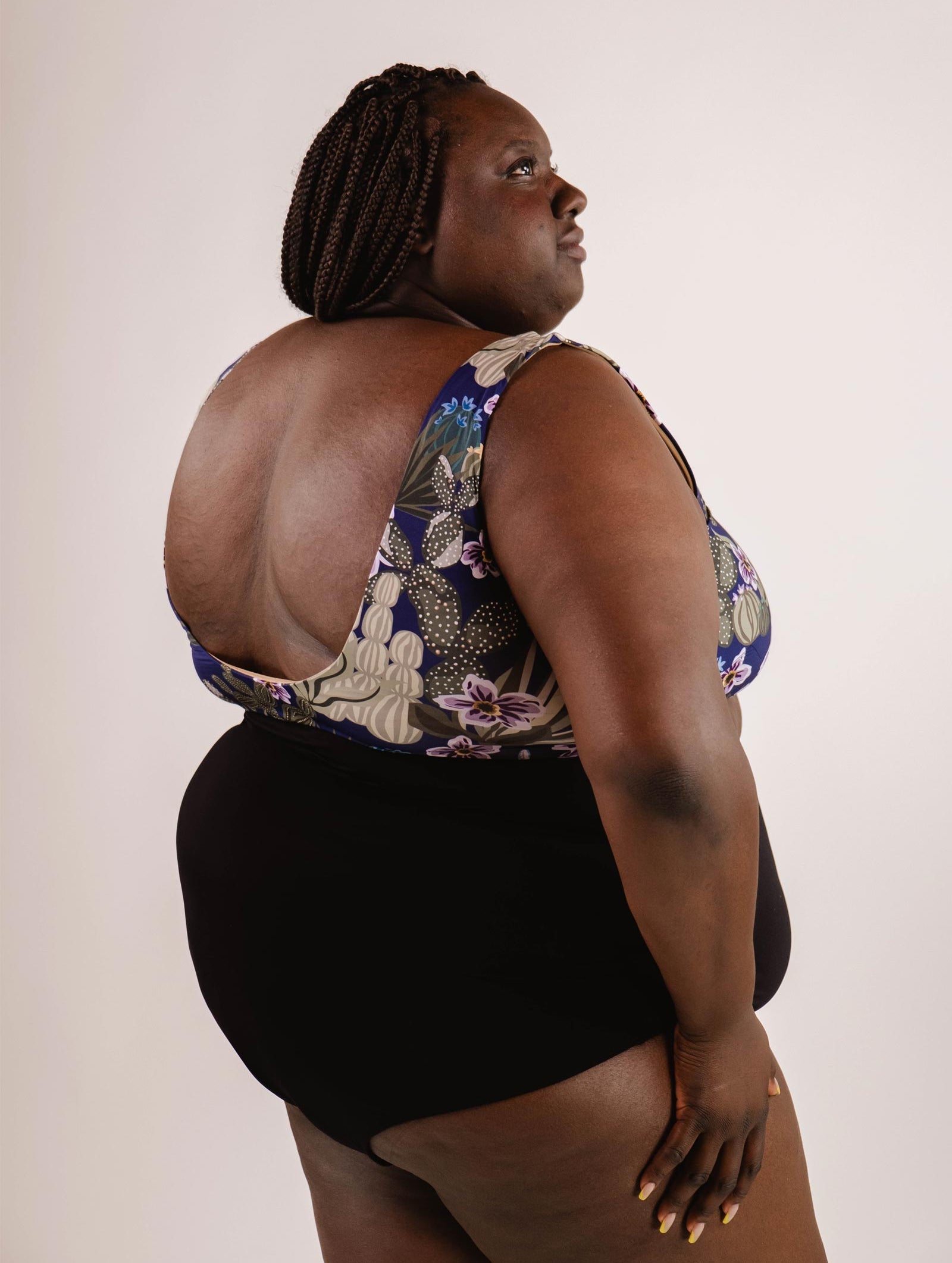 A person with braided hair wearing a floral-patterned swimsuit, featuring a Bermudes Black extra high waisted bikini bottom by Mimi & August, poses against a plain background.