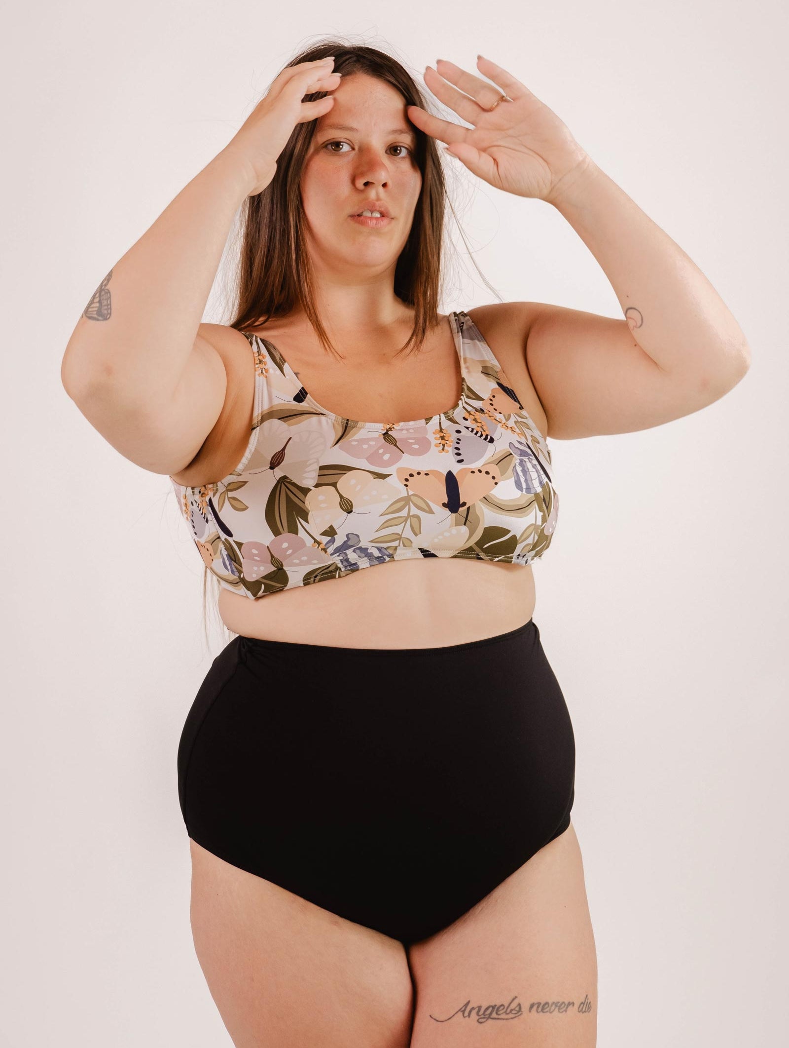 A woman with long hair wearing a floral bikini top and flattering, Mimi & August Bermudes Black extra high waisted bikini bottom stands against a neutral background, her hands raised near her head.